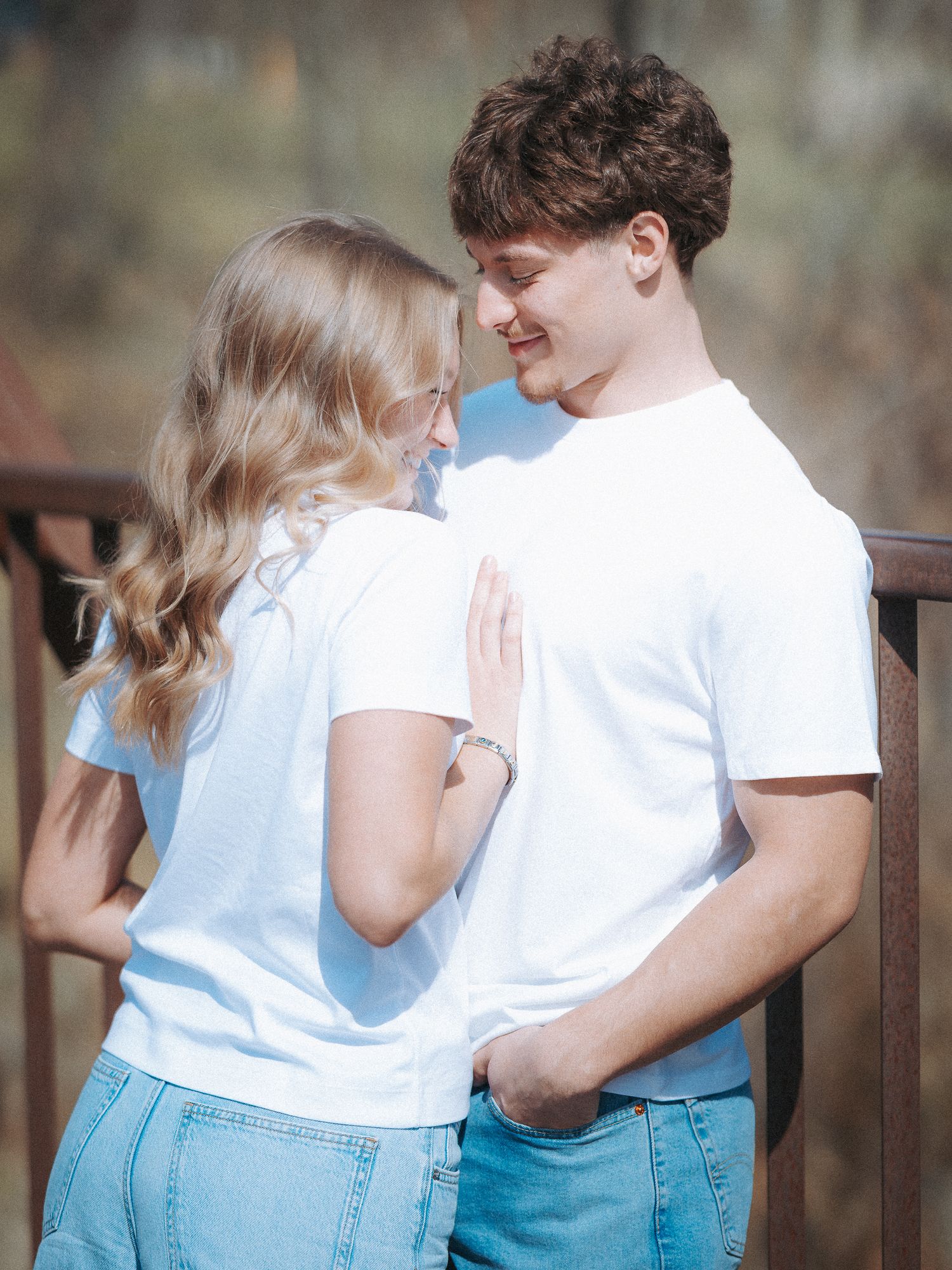A young couple in matching white t-shirts and jeans share a tender moment on a wooden bridge outdoors.
