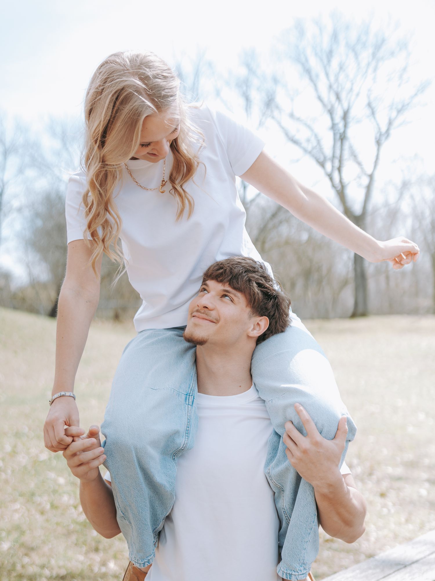 A young couple in white tees enjoy a playful piggyback ride outdoors in a sunny park setting.