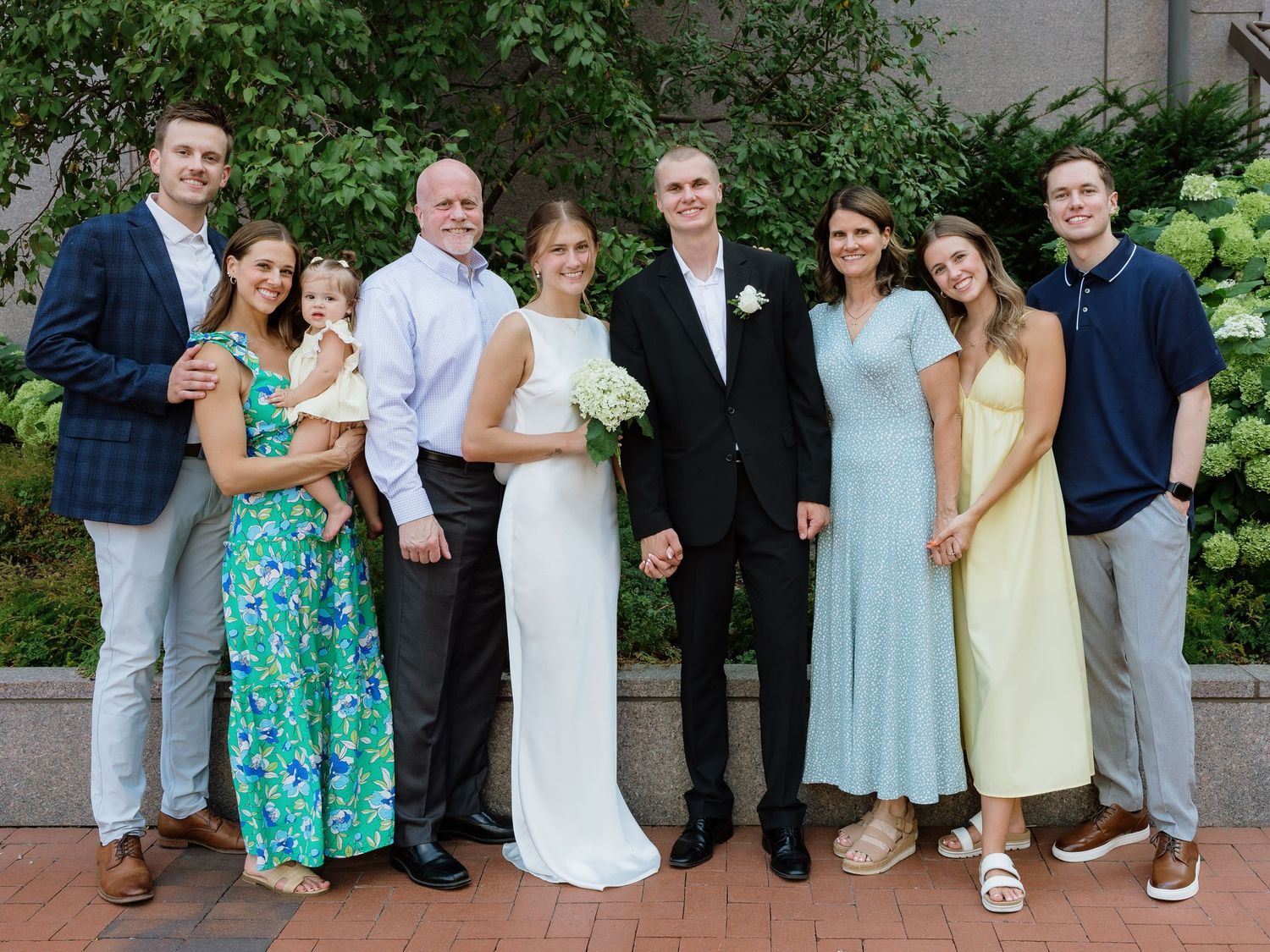 A wedding party of nine people poses outdoors, featuring a bride in white and groom in black suit surrounded by family.