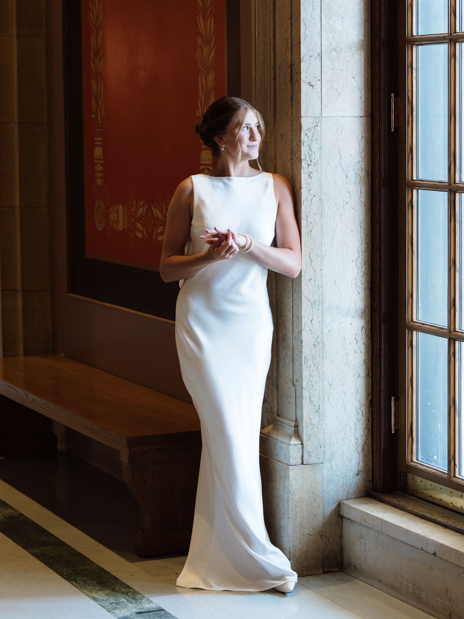 Bride in elegant white sleeveless gown standing by ornate window in grand marble hall, gazing thoughtfully to the side.