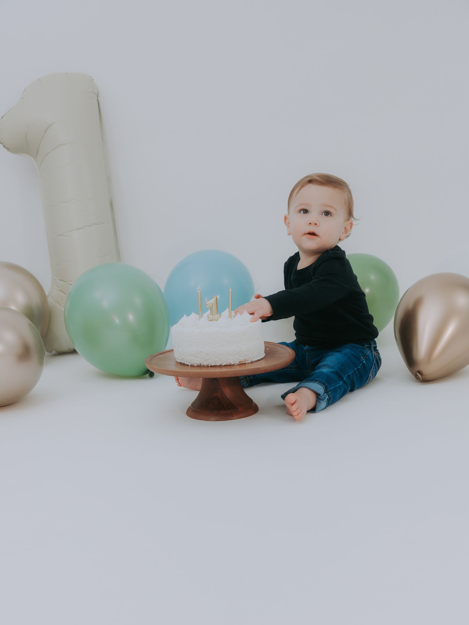 A baby boy in a black outfit sits beside a white first birthday cake surrounded by gold and green balloons.