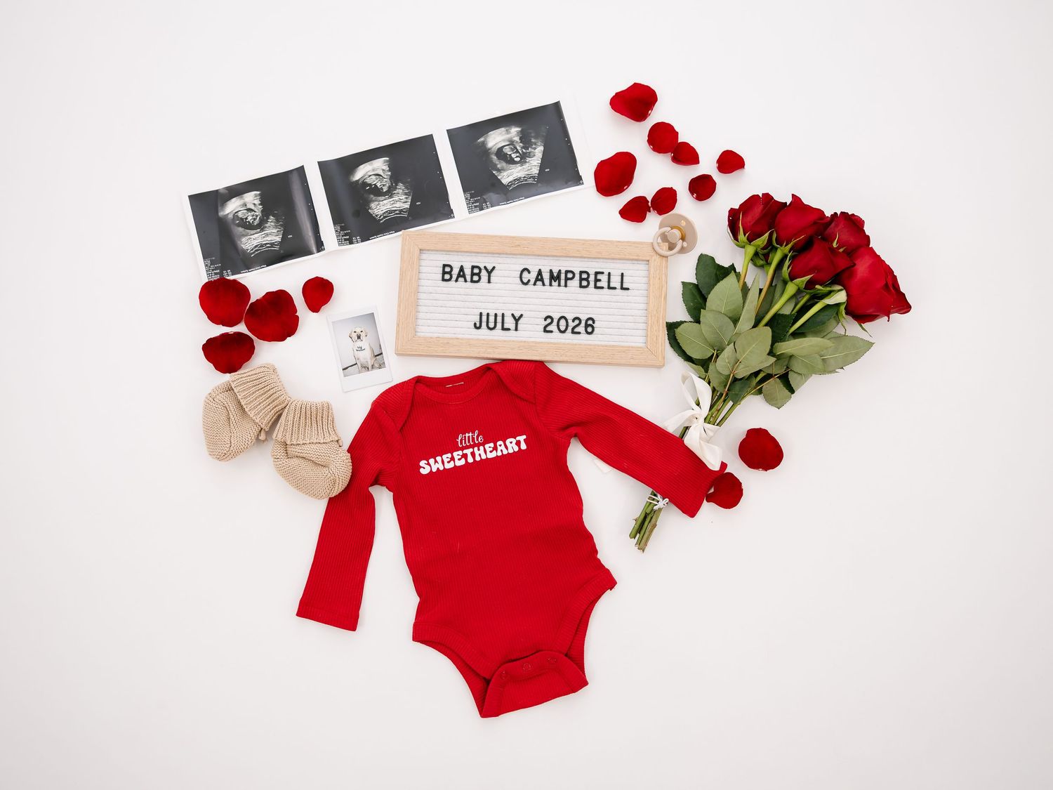 Baby announcement flatlay with red onesie, ultrasound photos, roses, and Baby Campbell July 2026 sign.