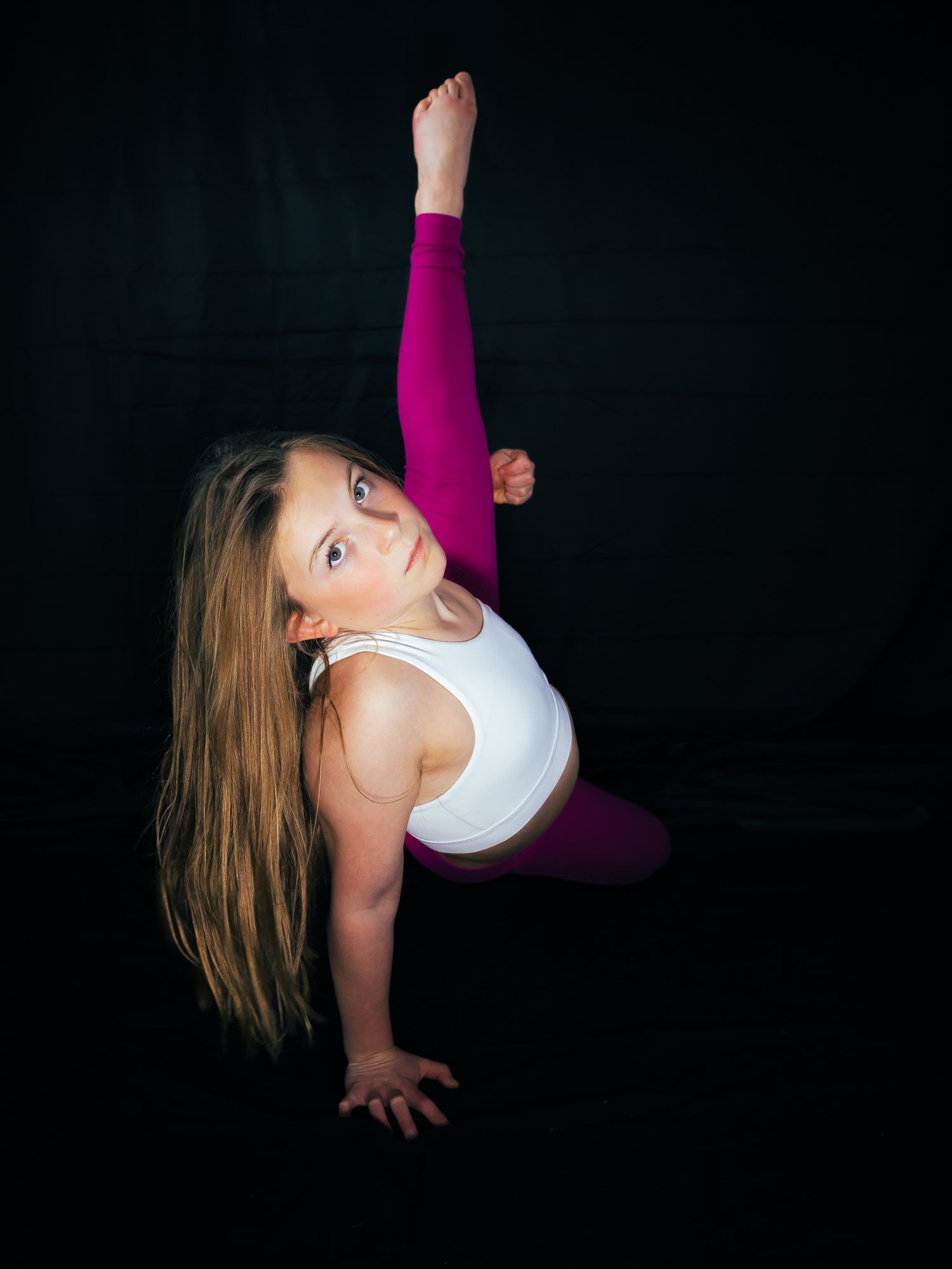 Young girl in white top and pink leggings performing a gymnastics pose against a dark black background.