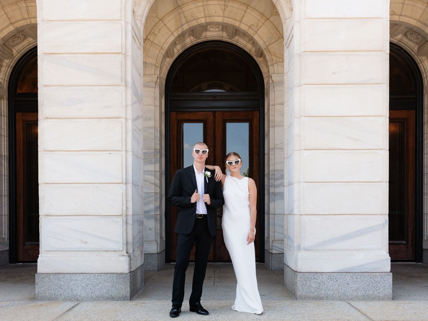 Elegant couple in wedding attire posing before ornate stone arched doorway.