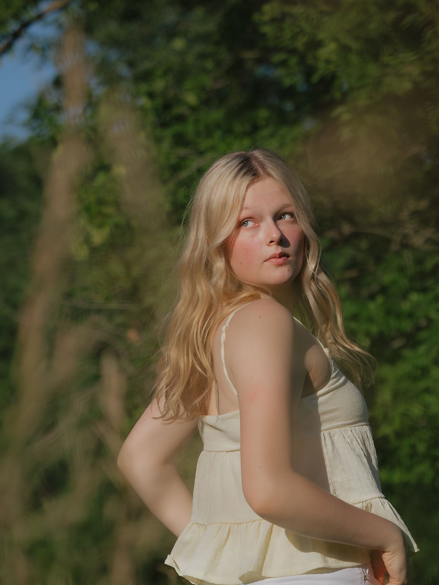 A young blonde woman in a yellow camisole gazes upward surrounded by lush green trees in golden light.