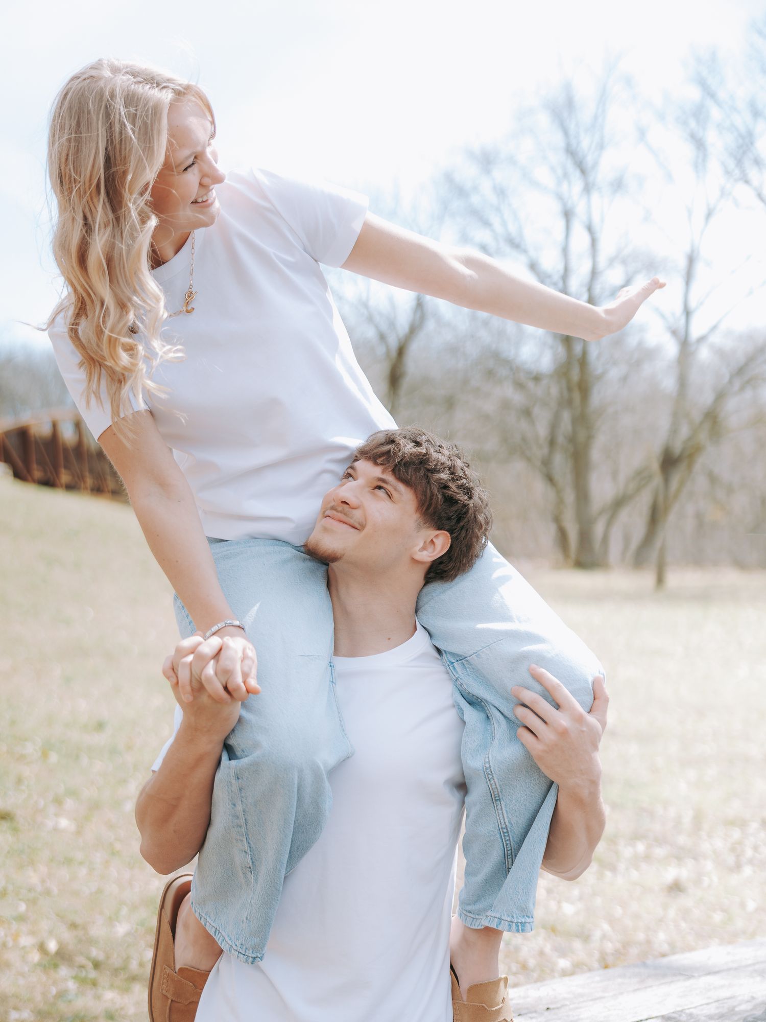 A smiling couple outdoors, woman sitting on mans shoulders with arms outstretched in a field.