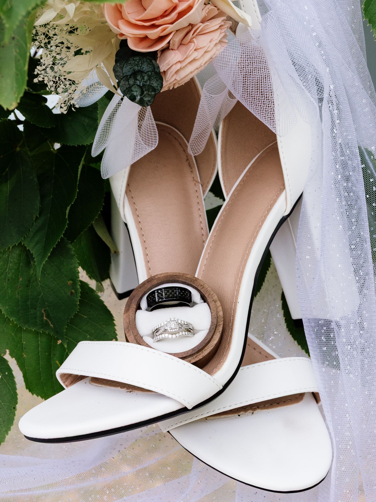White block heel bridal shoes with wedding rings displayed on floral bouquet background.