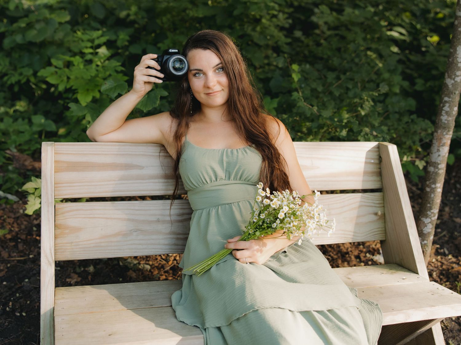 Young woman in green dress sitting on wooden bench with camera and wildflowers.