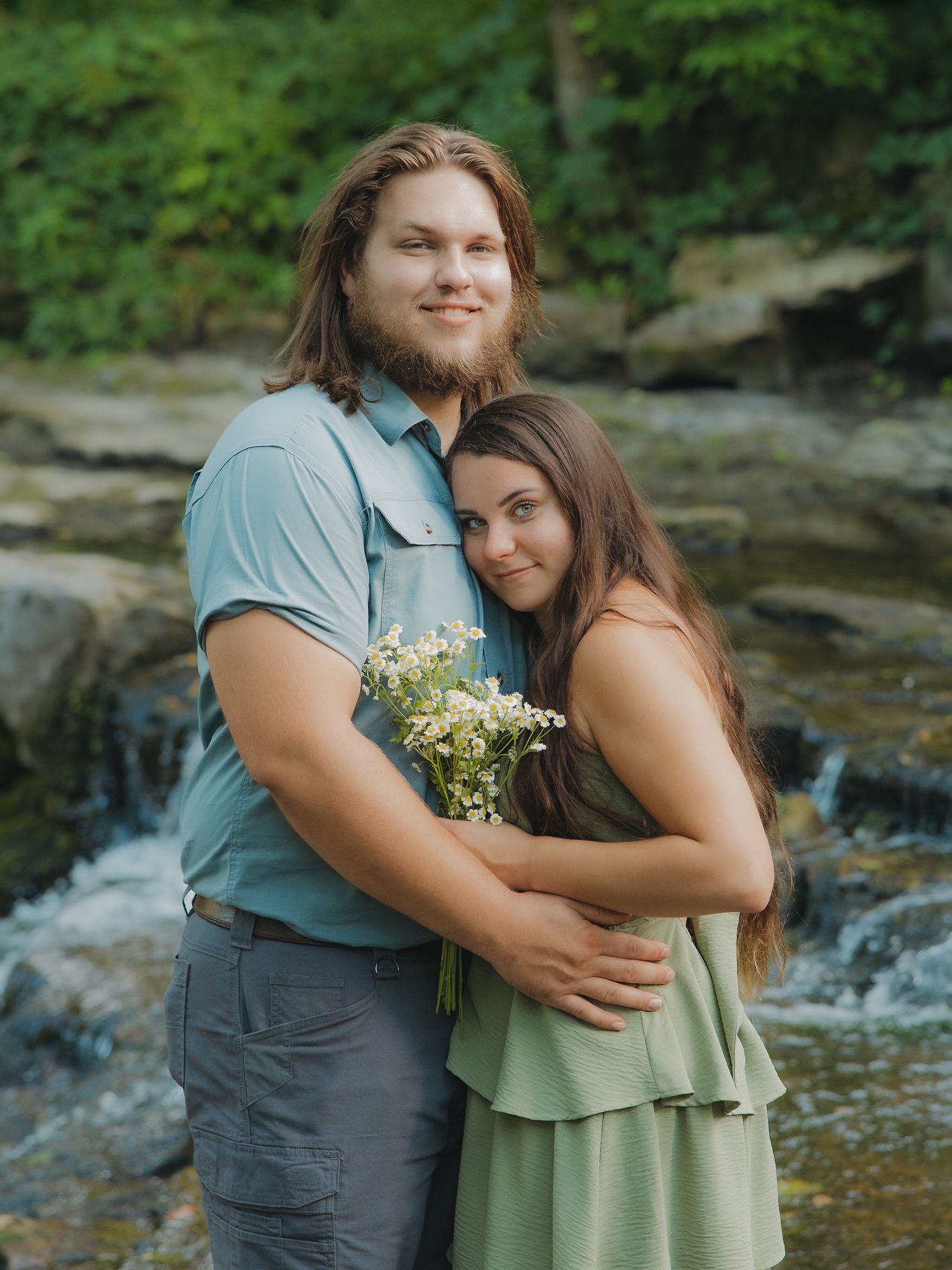 Couple posing by a rocky creek, woman holding wildflowers in a serene nature engagement portrait.