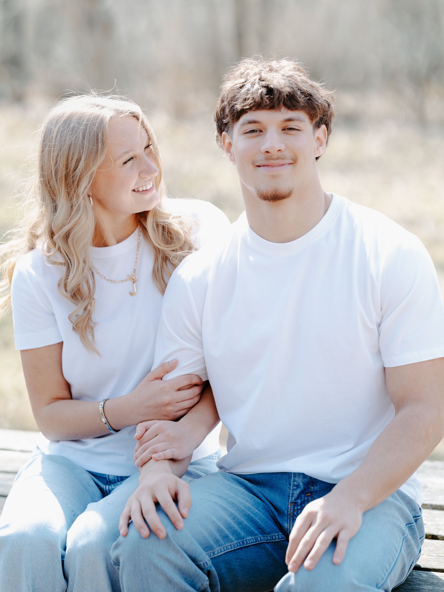 A young couple in matching white t-shirts and jeans sit closely together outdoors, smiling warmly in a bright natural setting.