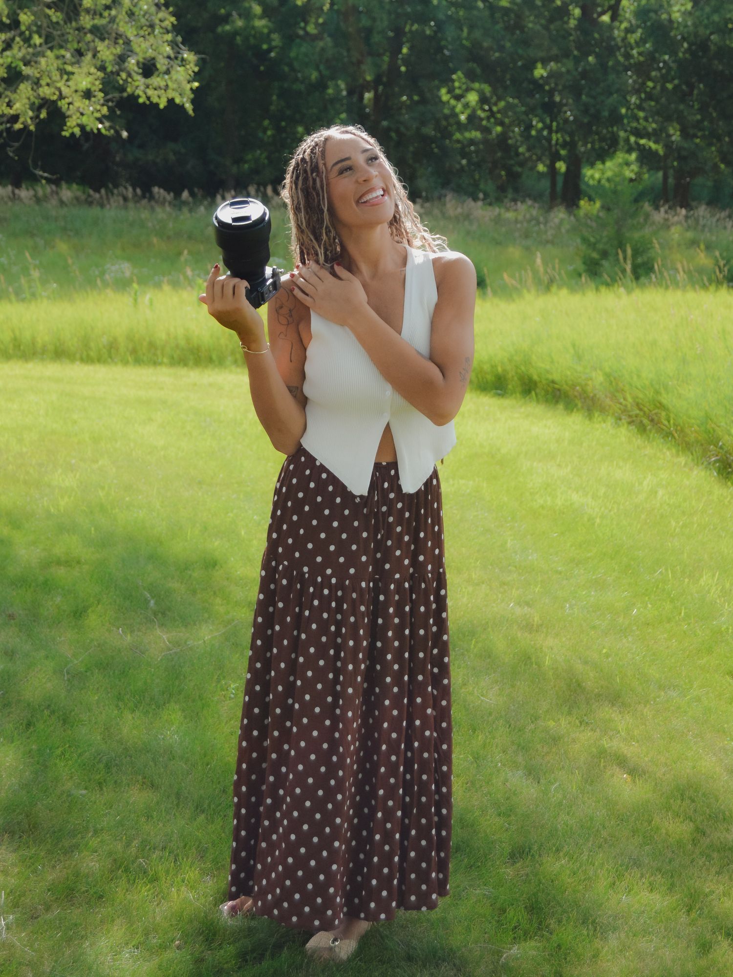 Smiling woman in white vest and polka dot skirt holding a camera in a sunny green park.