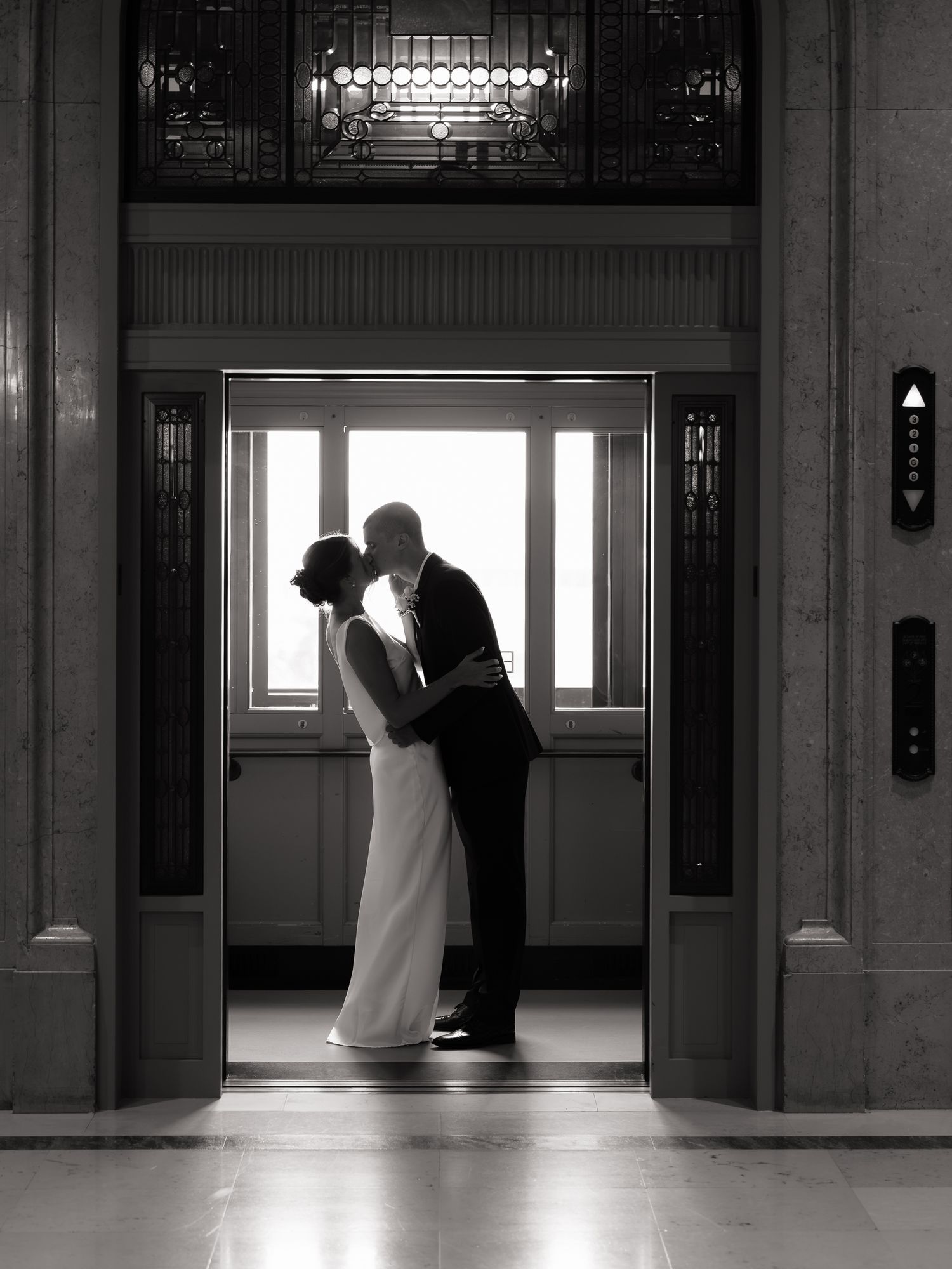 Black and white photo of bride and groom sharing a kiss in elegant building elevator doorway.