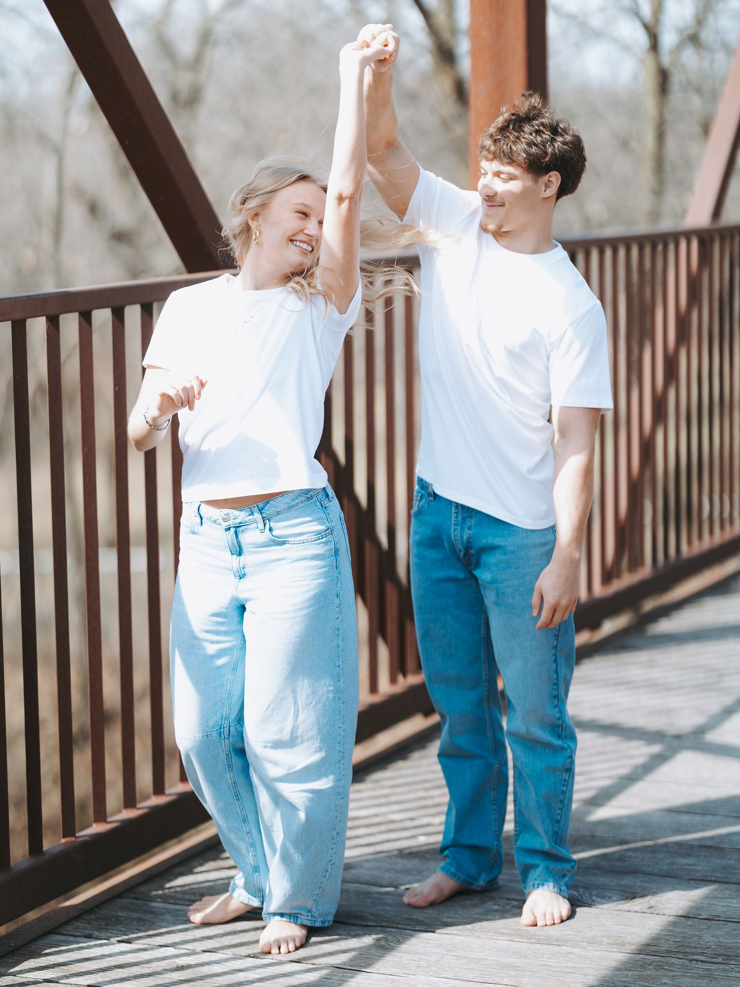 Happy couple in white tees and jeans dancing barefoot on a sunny wooden bridge.