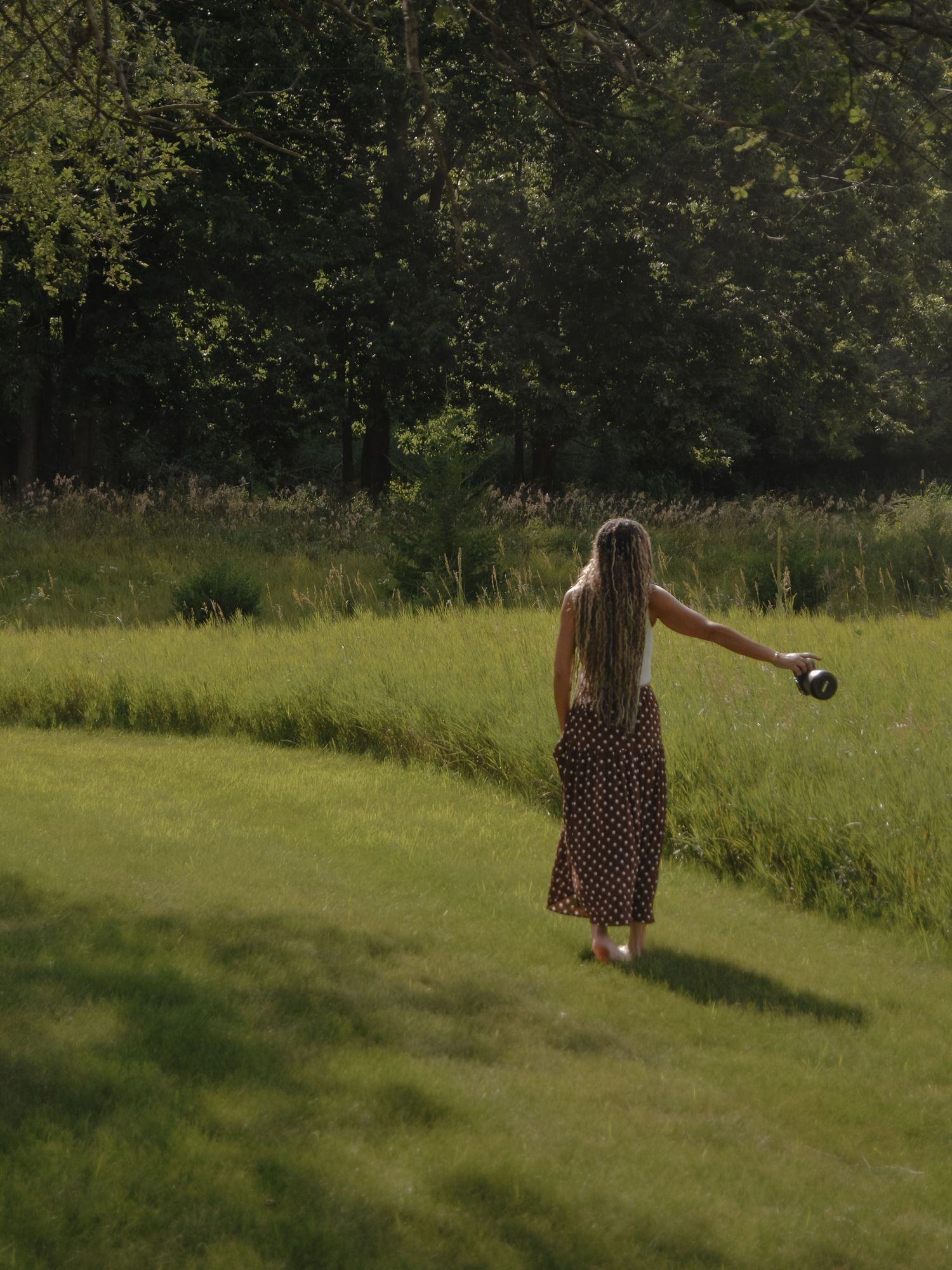 Woman in floral maxi dress strolling through sunlit green meadow holding a camera at golden hour.
