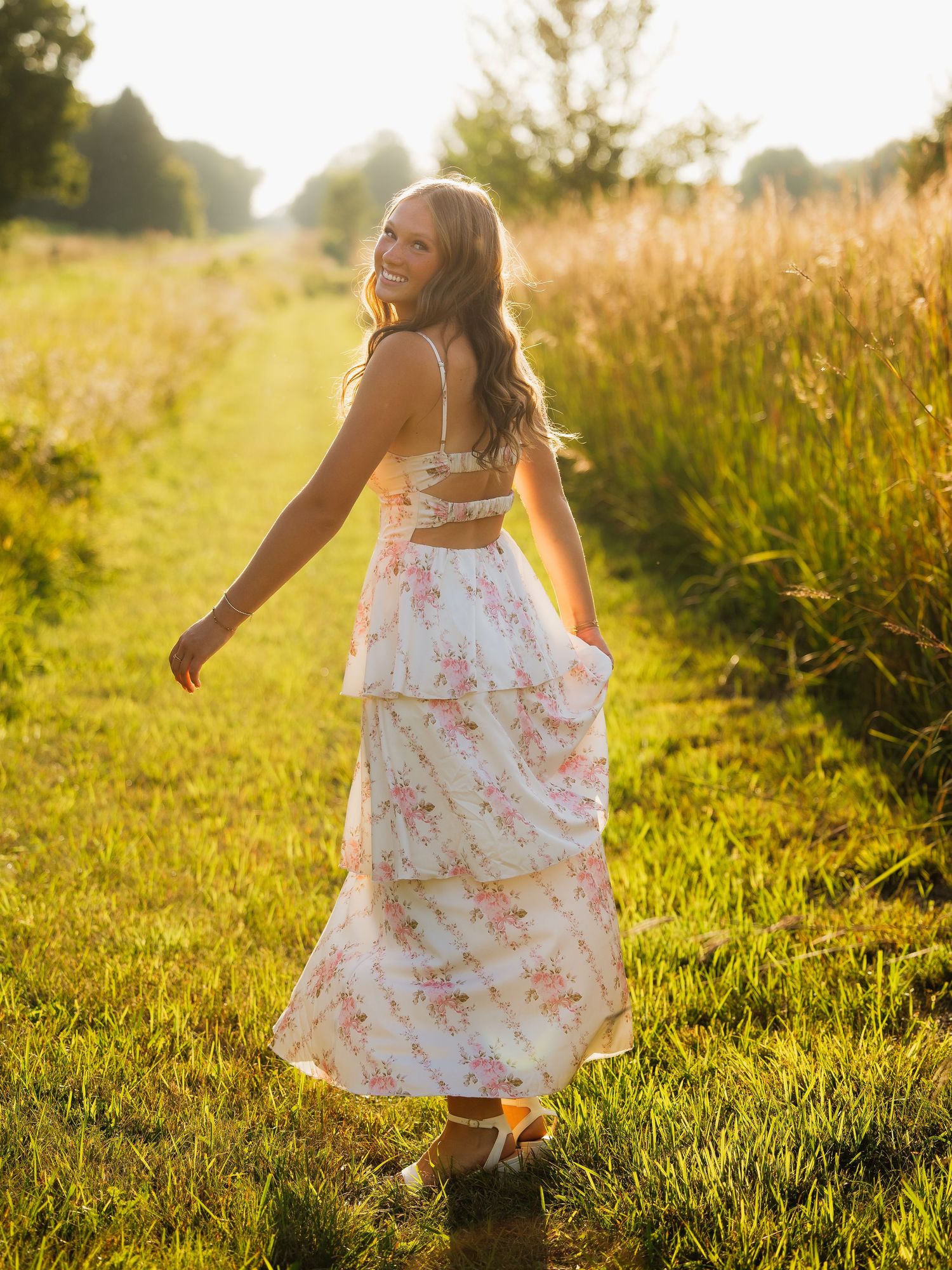 Young woman in floral maxi dress walking through sunny green field, smiling over shoulder in golden hour light.