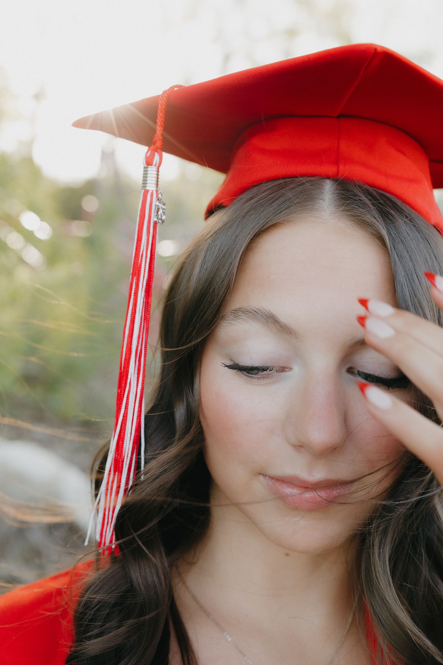A brunette woman in a red graduation cap and gown poses outdoors in soft light.