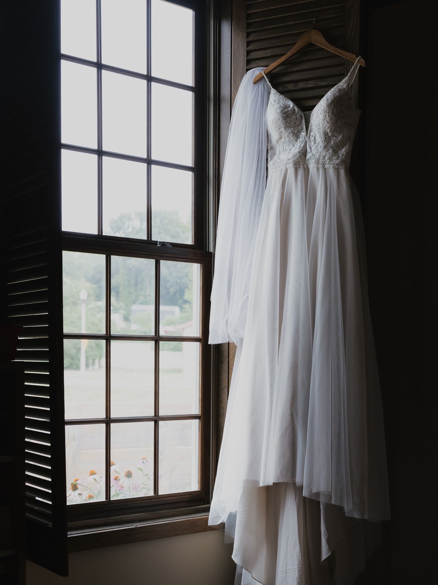 Elegant white lace wedding dress with veil hanging on wooden hanger in window.