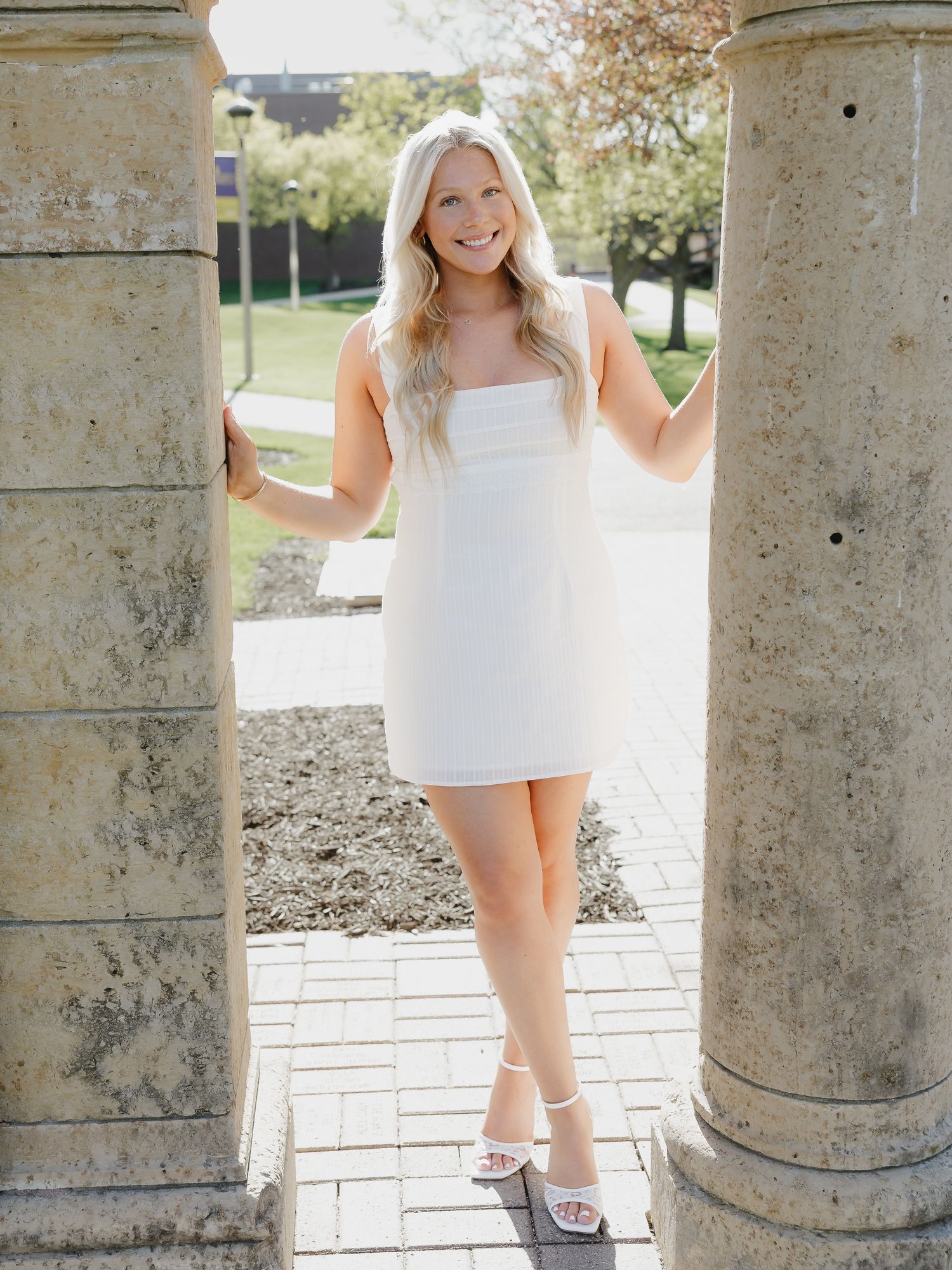 Blonde woman in white mini dress posing between stone columns on sunny day.