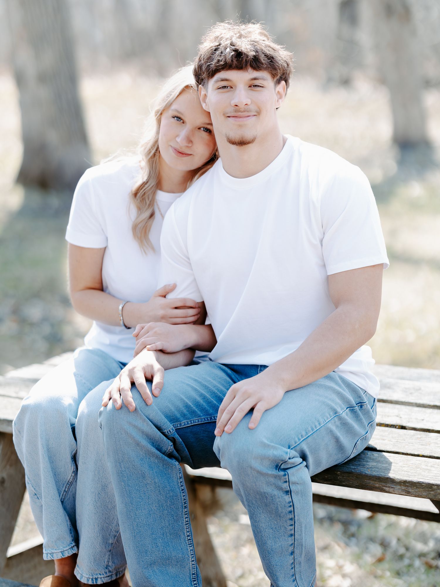 A young couple in matching white tees and jeans sit closely on a wooden bench outdoors, smiling warmly.