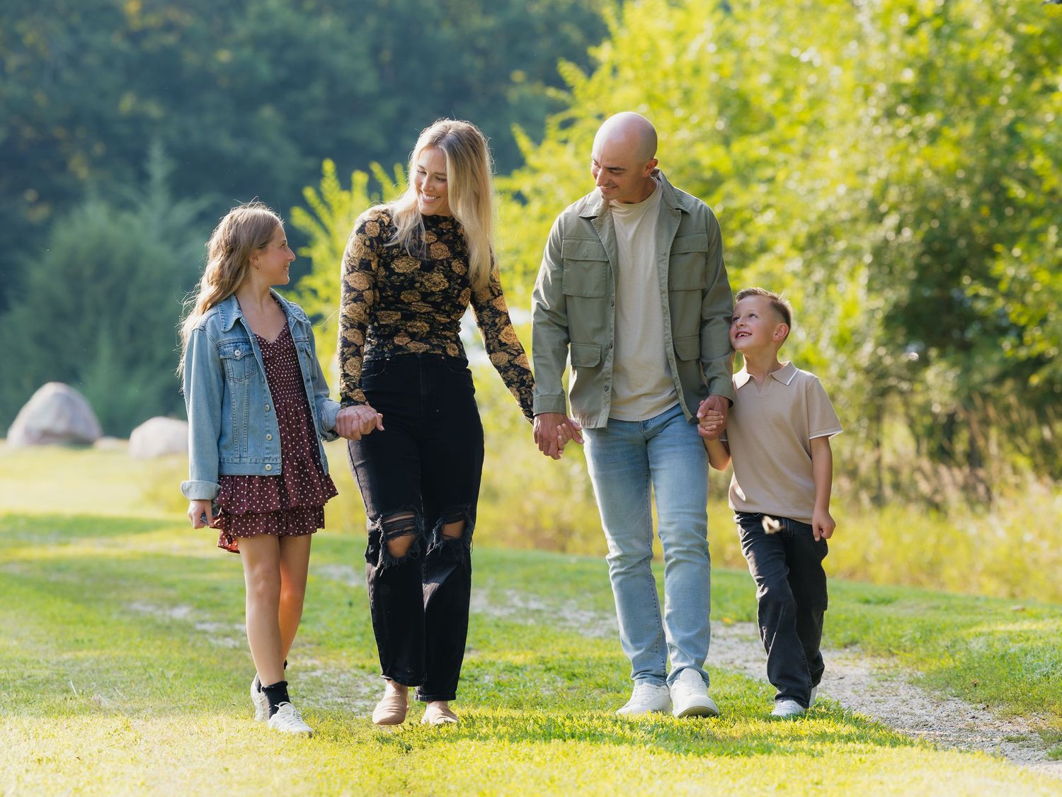 Family of four walking hand-in-hand on a sunny park path, smiling and enjoying quality time together outdoors.