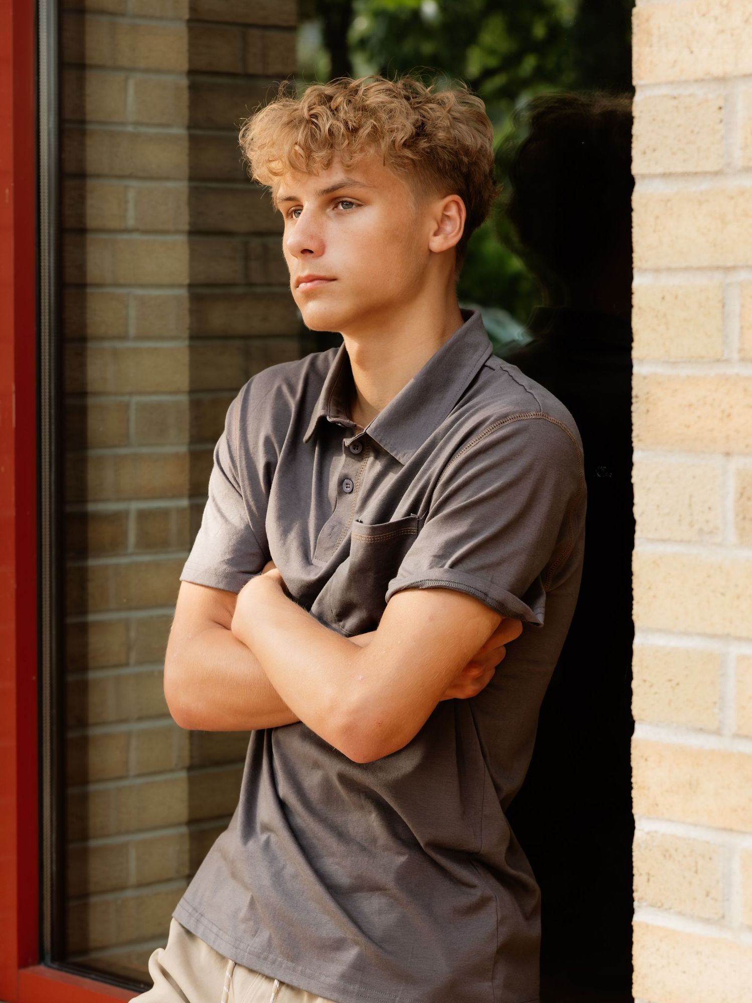 A teenage boy with curly blonde hair stands with arms crossed, wearing a gray polo shirt, leaning against a brick wall.