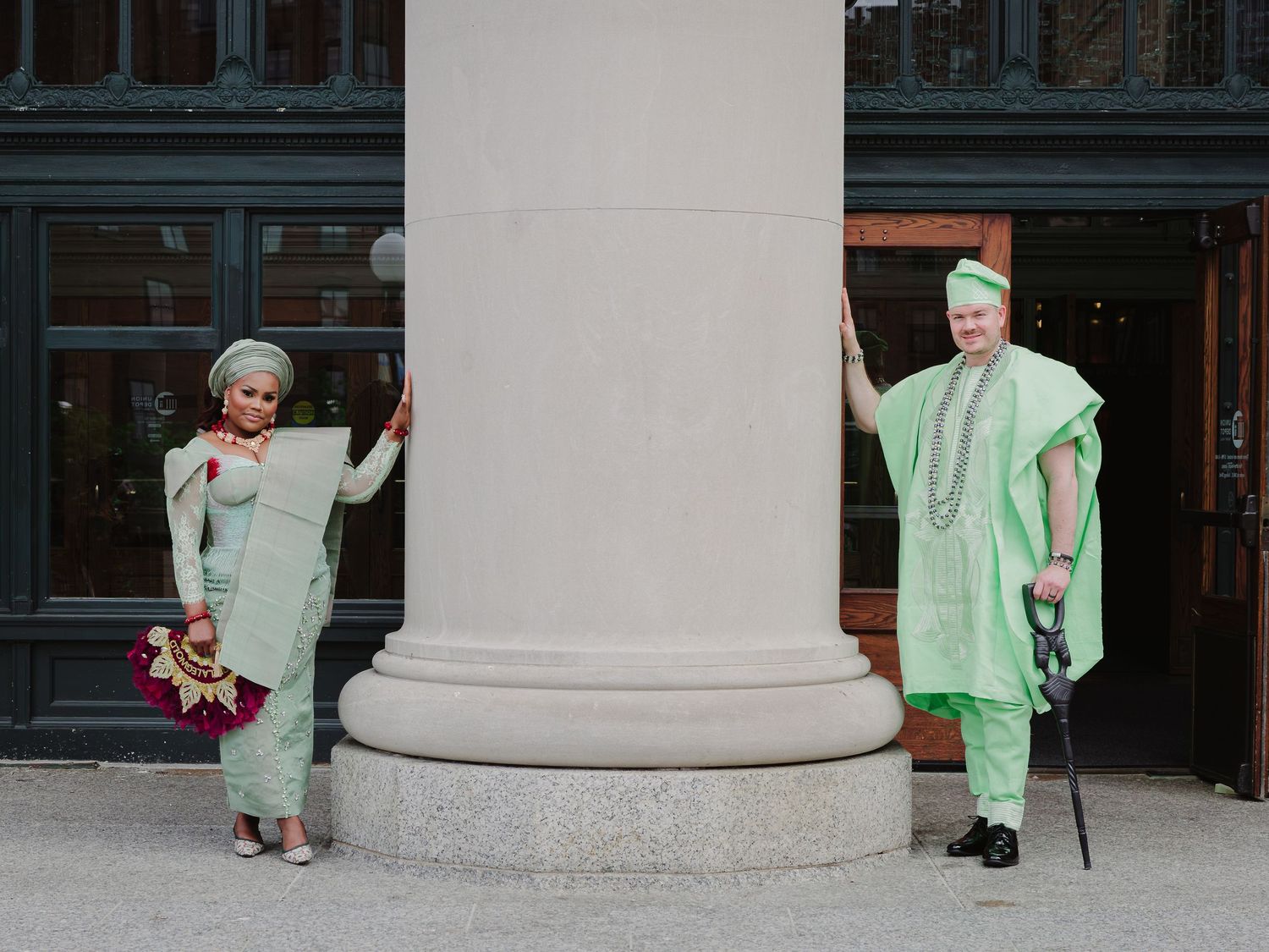 A couple in matching mint green traditional attire poses beside a column outside a building.