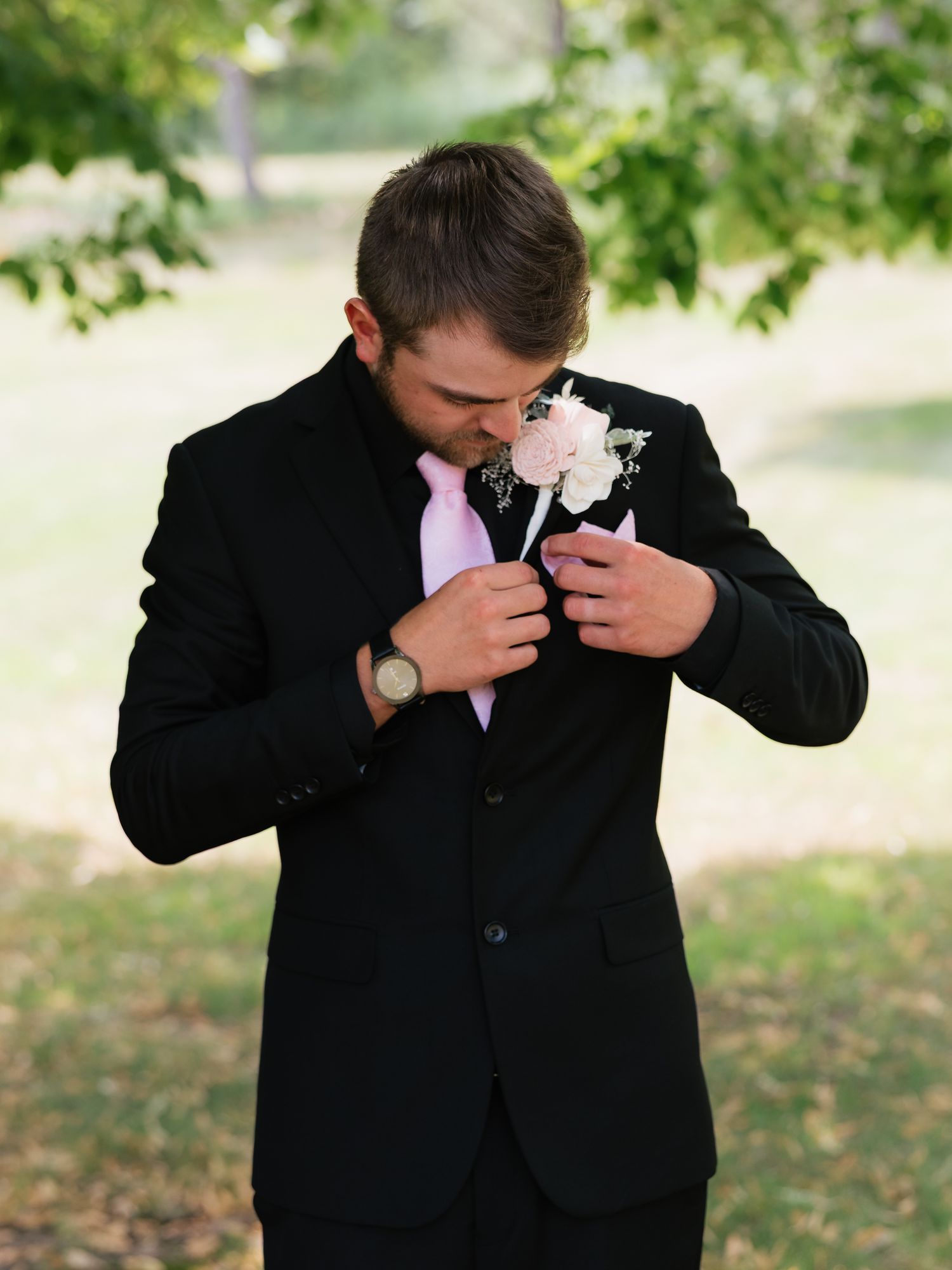 Groom in black suit adjusting his floral boutonniere outdoors under green trees in Minnesota