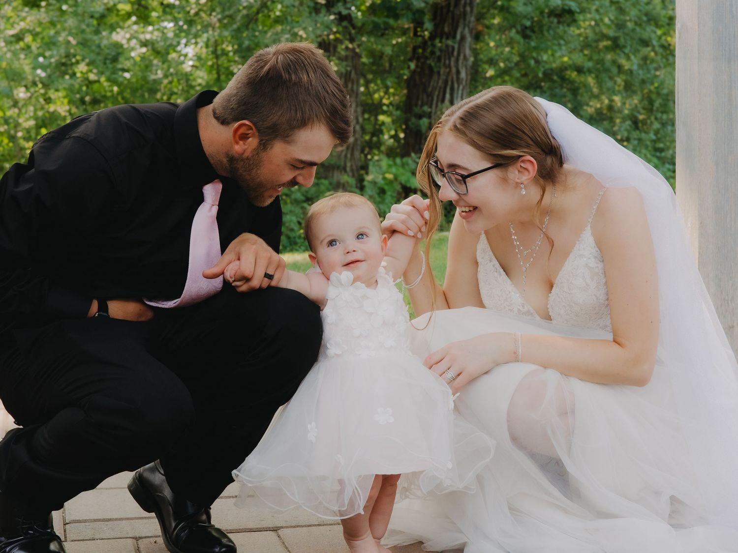Bride and groom crouching to interact with a baby girl in white dress.