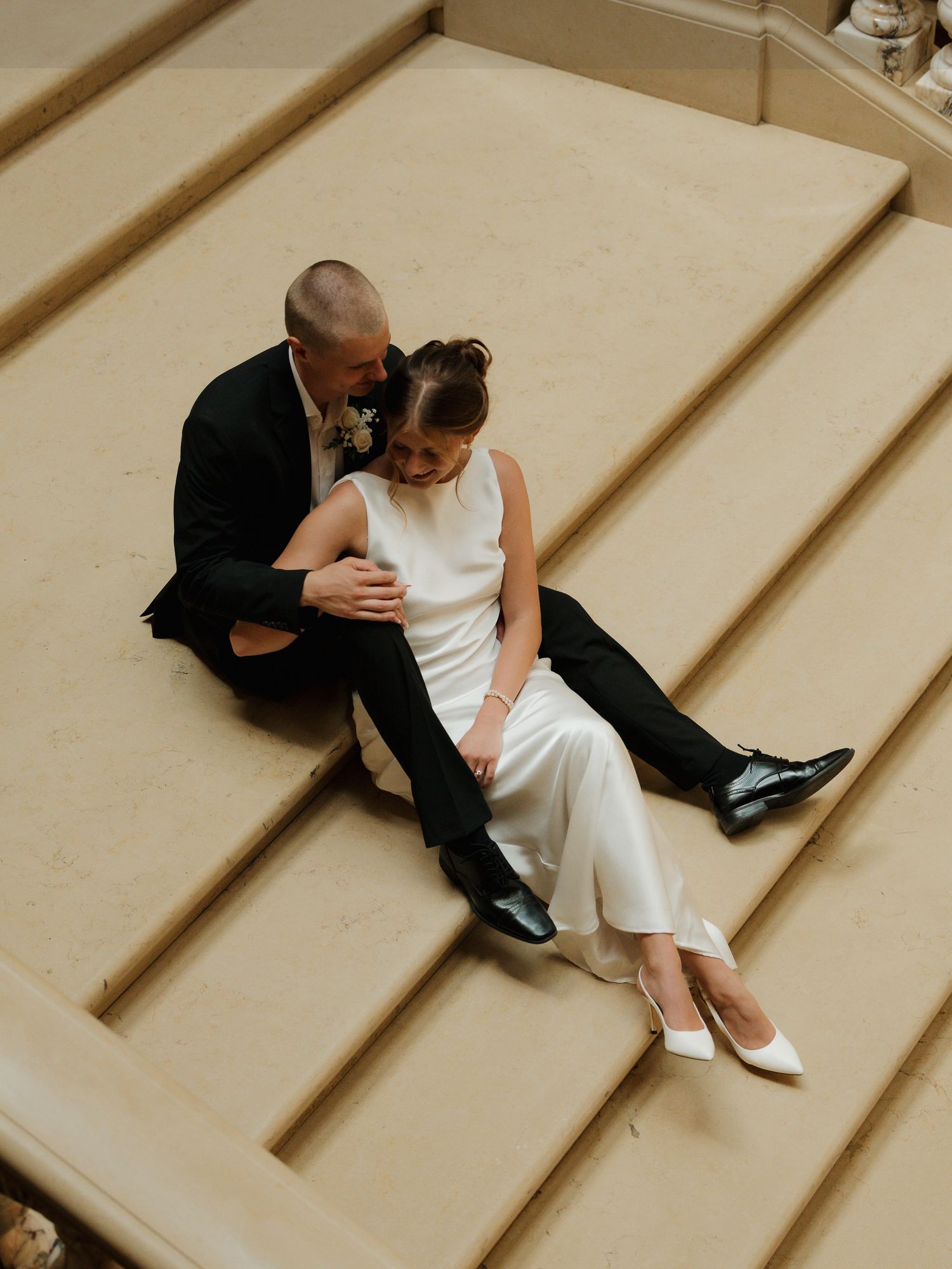 Bride and groom sitting on grand stone steps, she in white jumpsuit and heels, he in black suit, sharing an intimate moment.