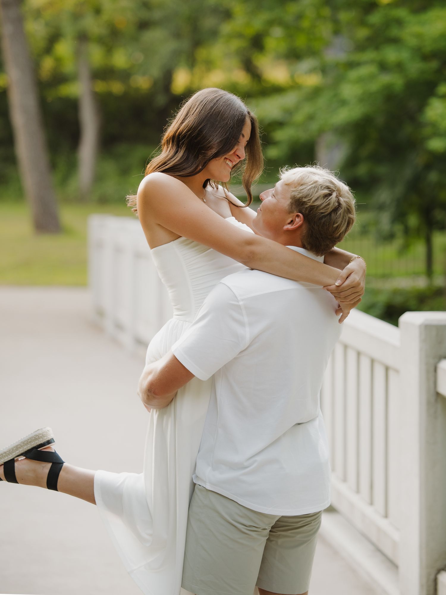Couple embracing on a white bridge in a lush green park during a summer engagement session.