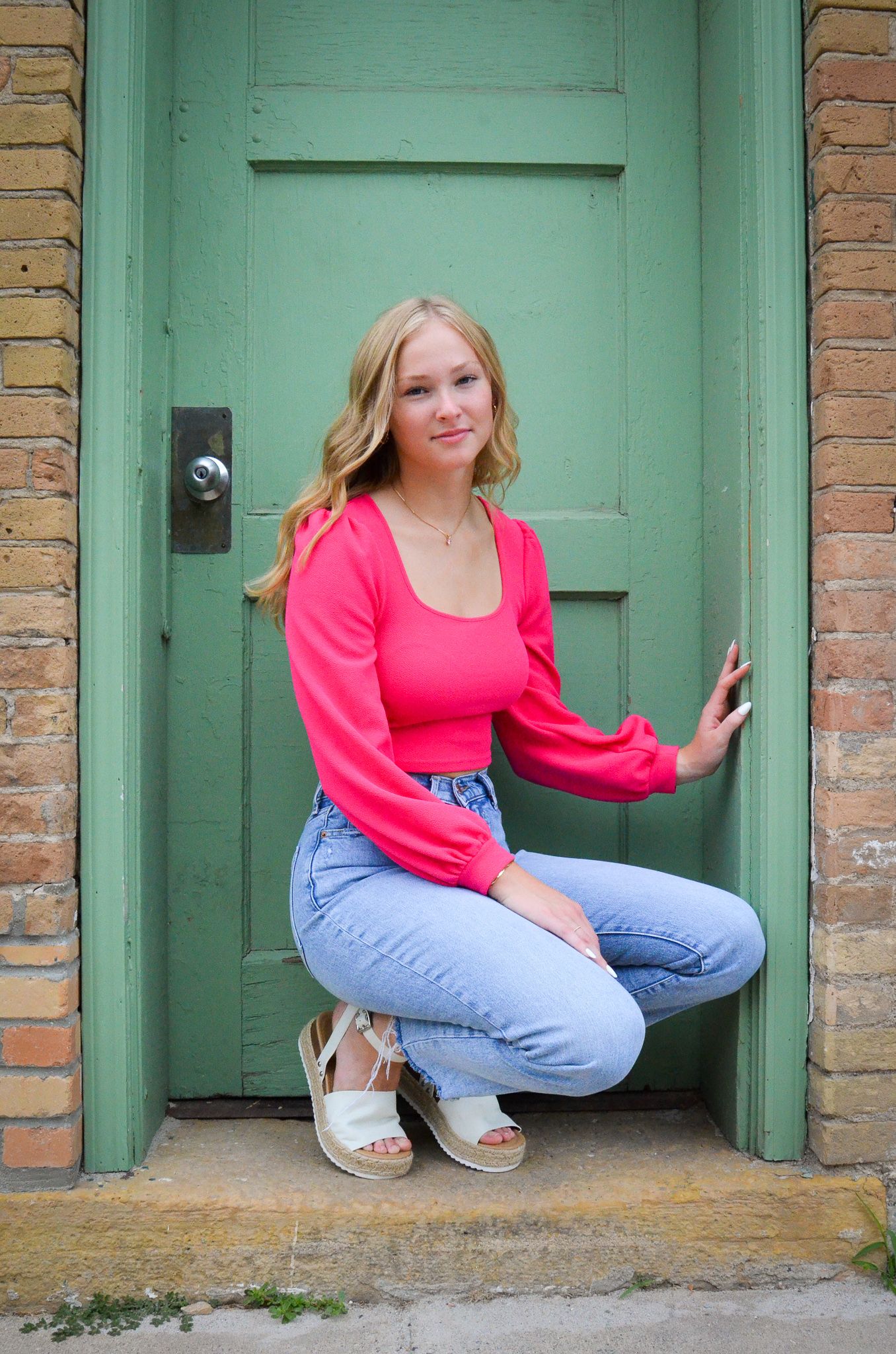 Blonde teenage girl in pink top and jeans posing by a green door for senior photos.
