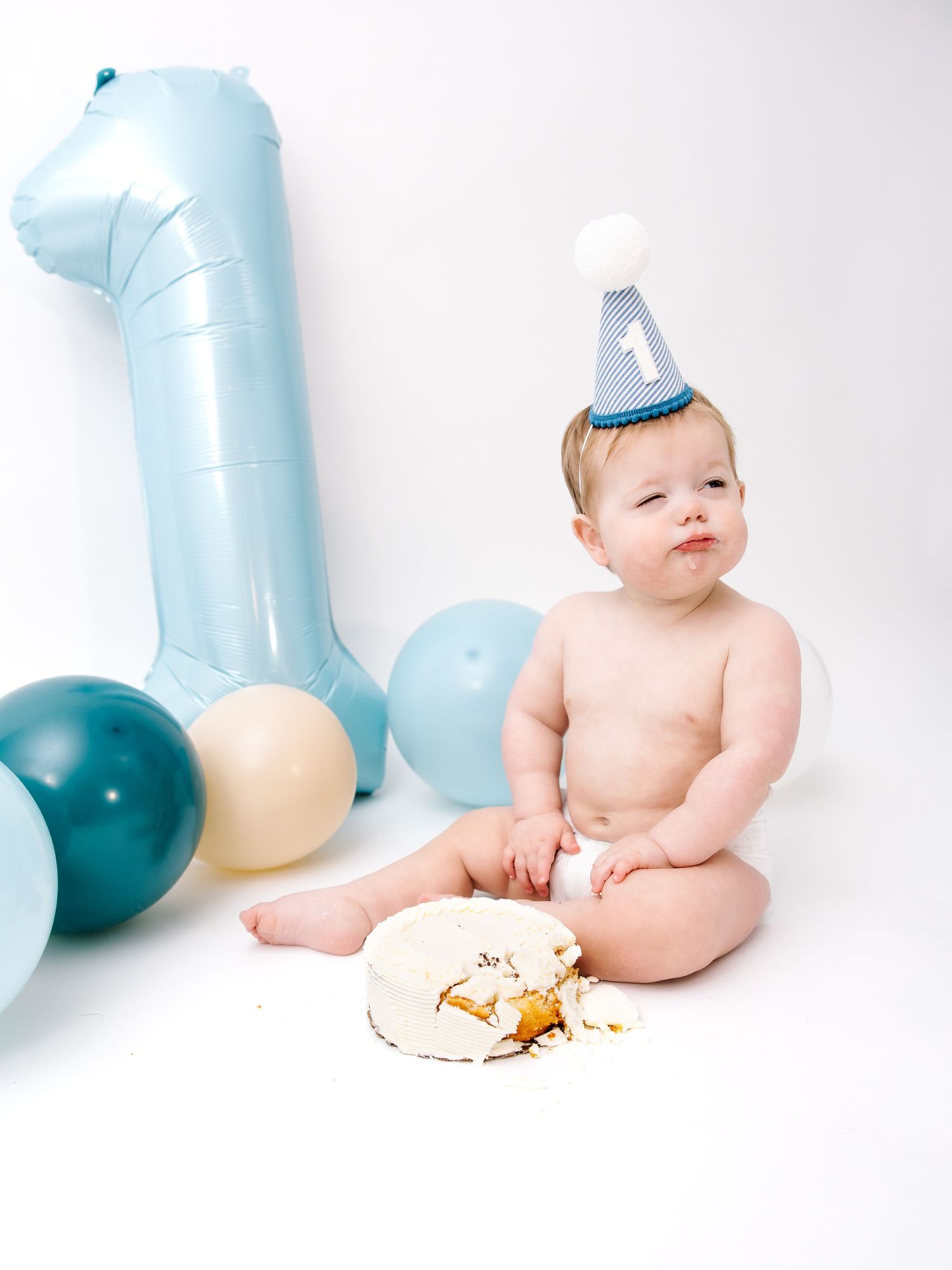 Baby's first birthday cake smash with blue number one balloon, party hat, and colorful balloons on white background.