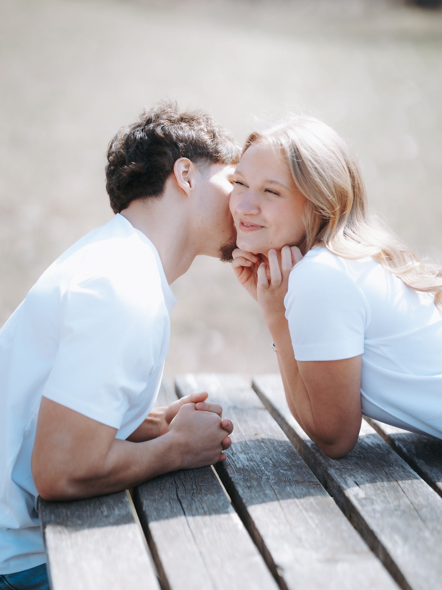 A young couple in matching white t-shirts sharing a tender kiss on a wooden bench outdoors in soft sunlight.