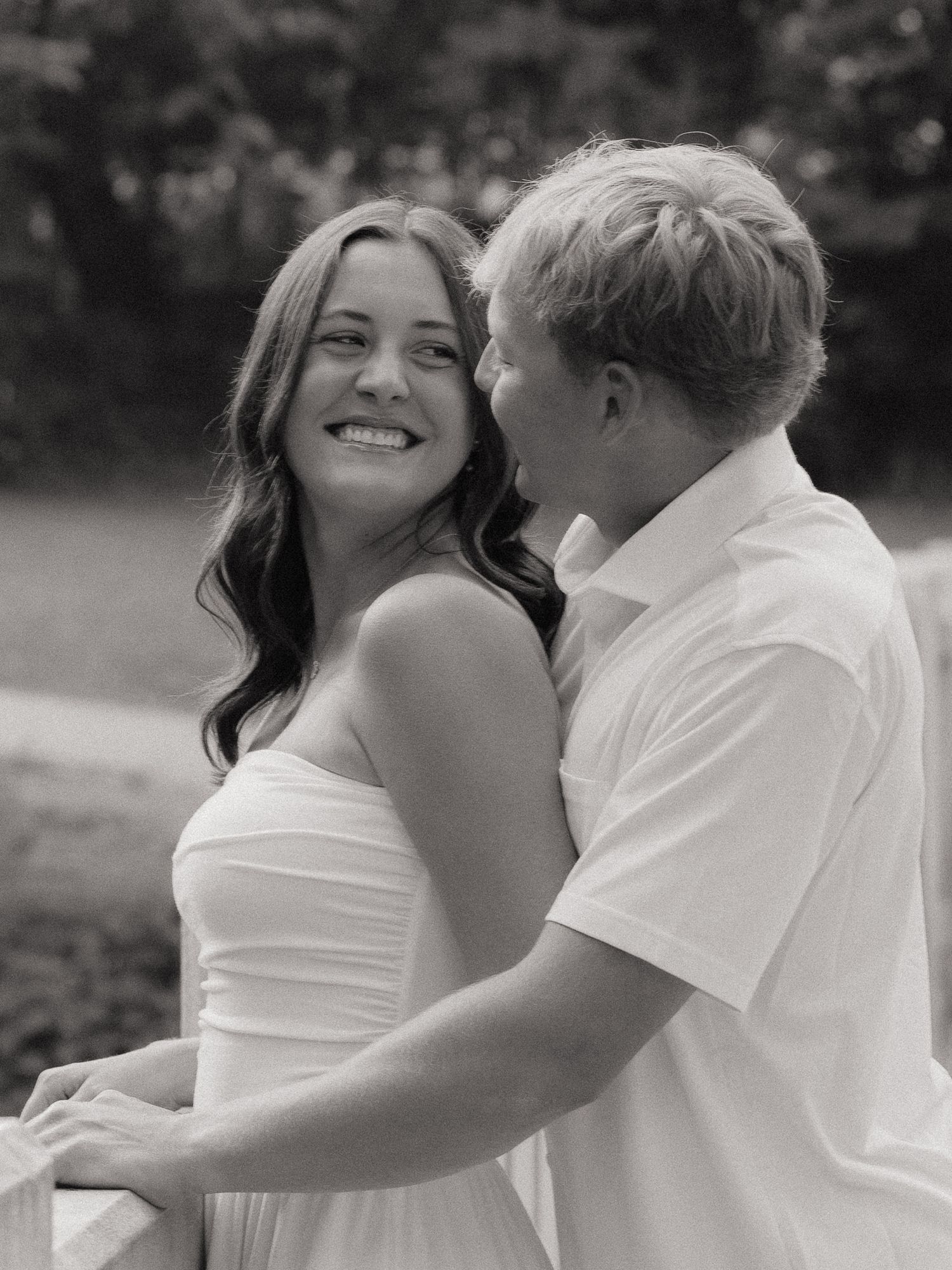Black and white photo of a smiling couple embracing outdoors, woman in strapless dress, man in white shirt.