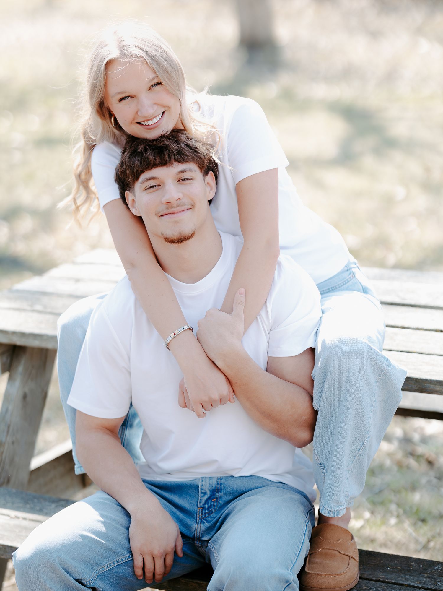 Smiling couple in white shirts and jeans posing on a wooden bench in a sunny park.