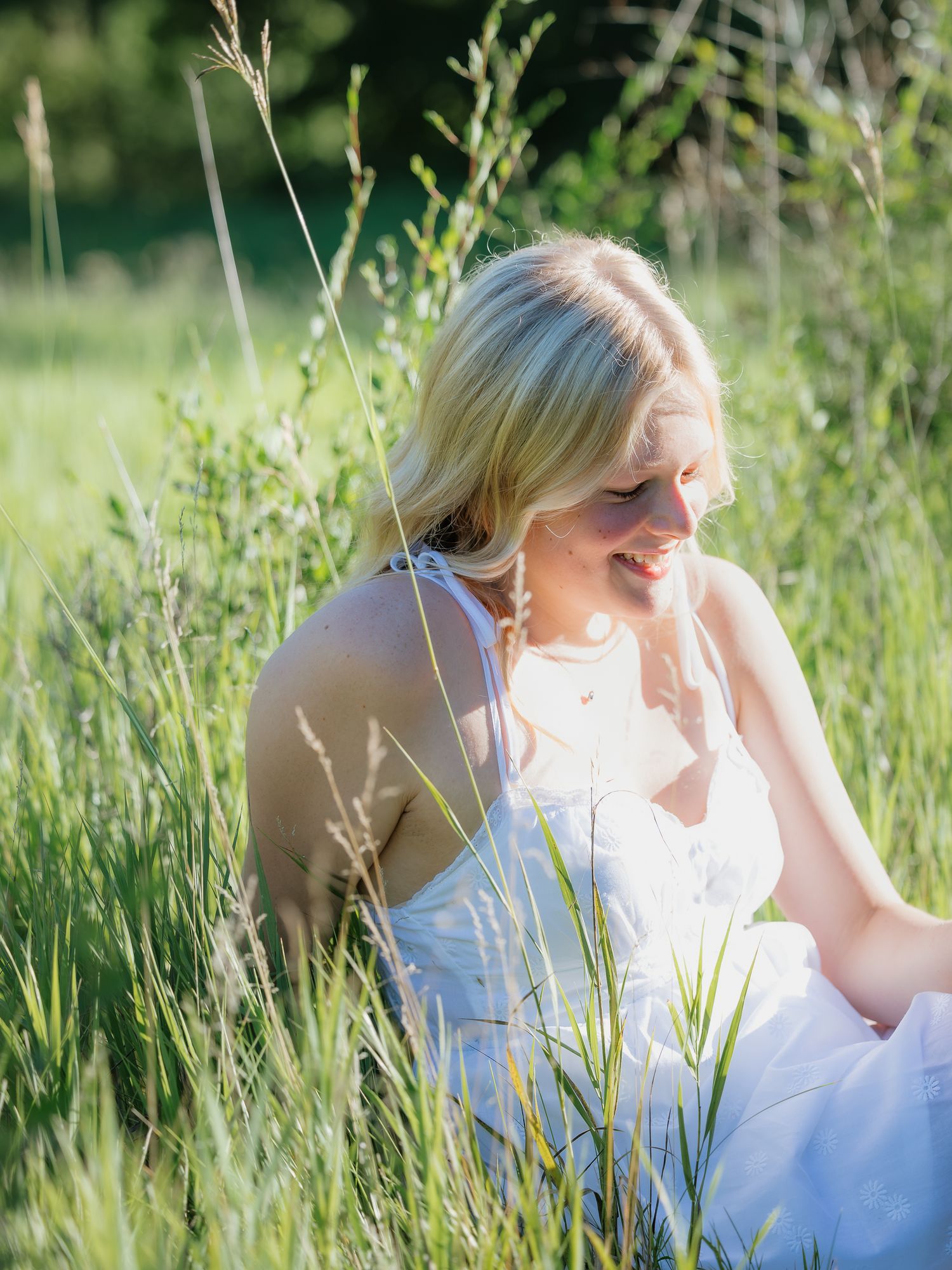 Blonde woman in white dress sitting peacefully in tall green meadow grass.