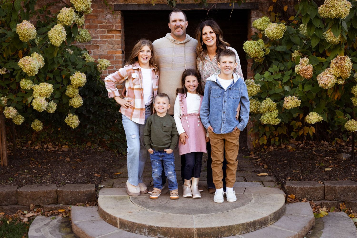 A family of seven smiles together on stone steps framed by blooming hydrangea bushes.