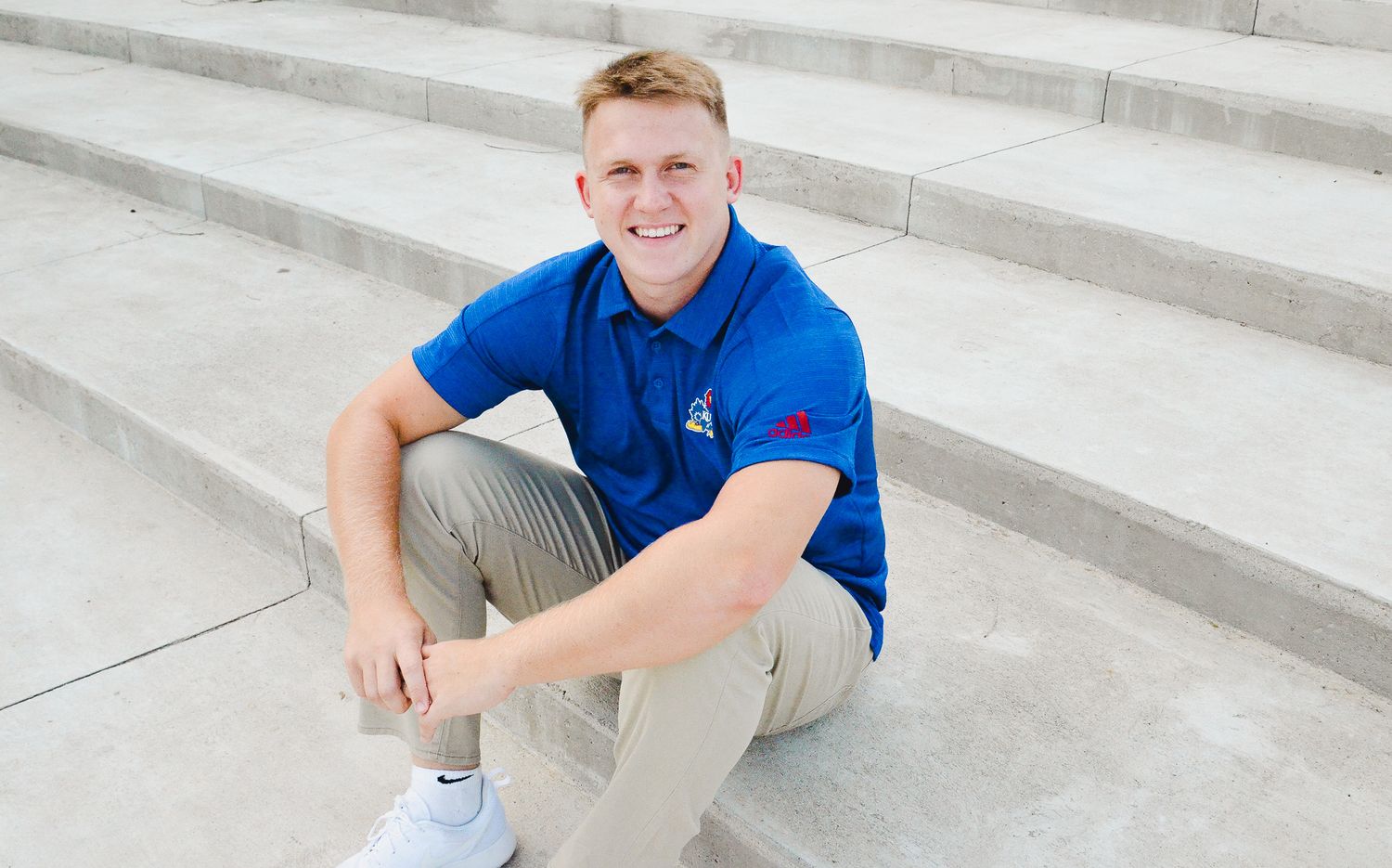 Smiling young man in blue polo shirt sitting on concrete steps outdoors.