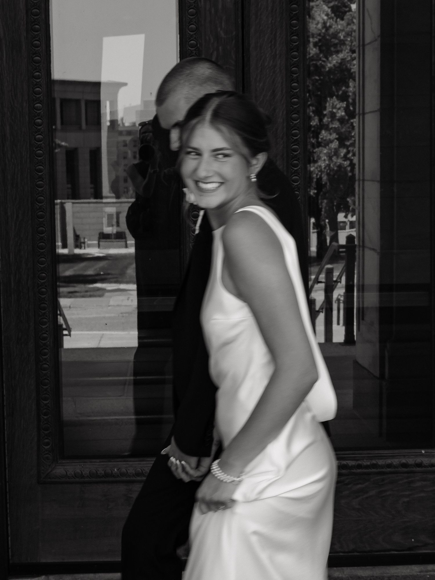 A bride in a white satin gown smiles over her shoulder while embracing her groom outdoors.