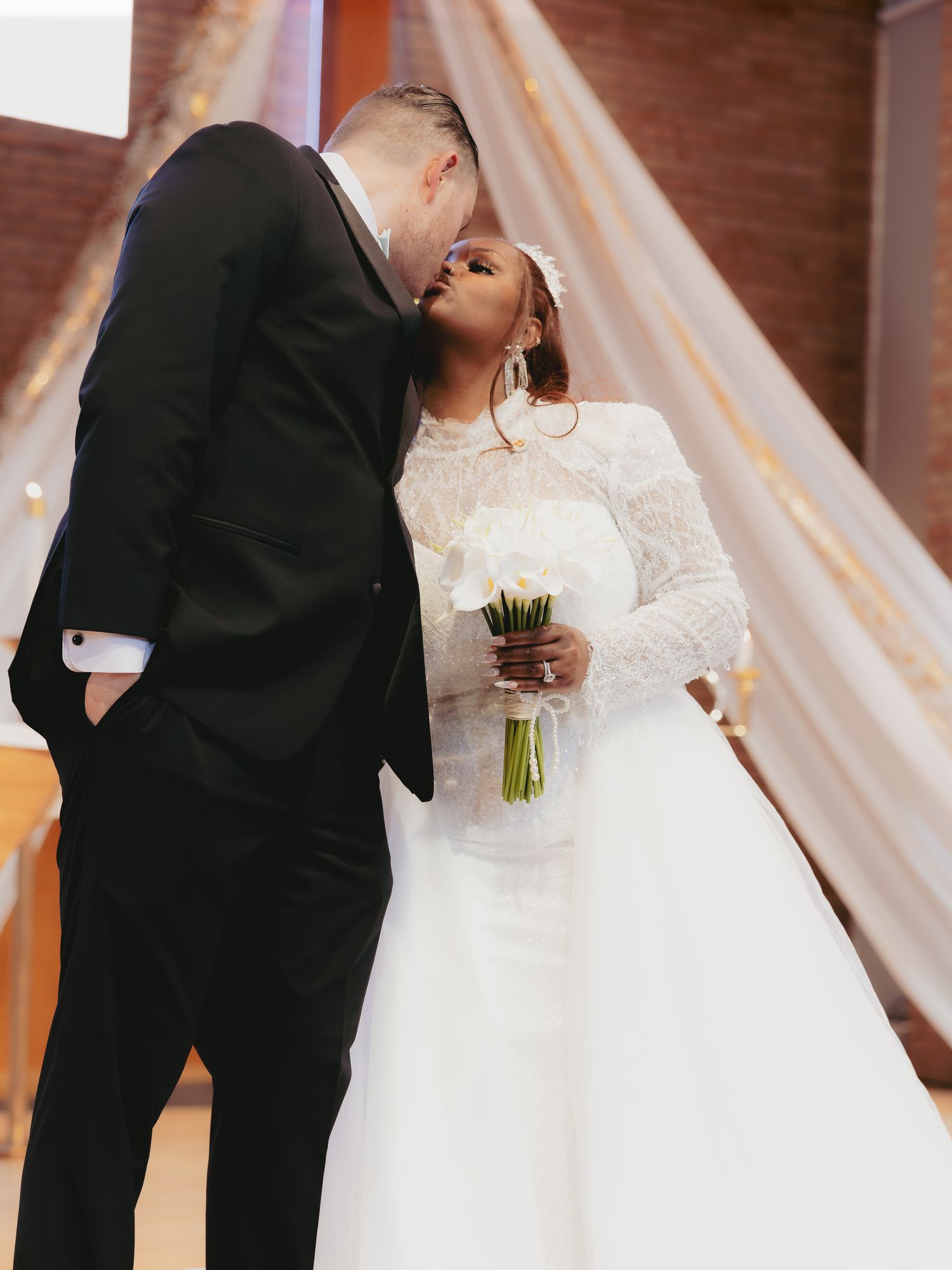 Interracial couple sharing a kiss at the altar, bride in lace gown holding white bouquet.