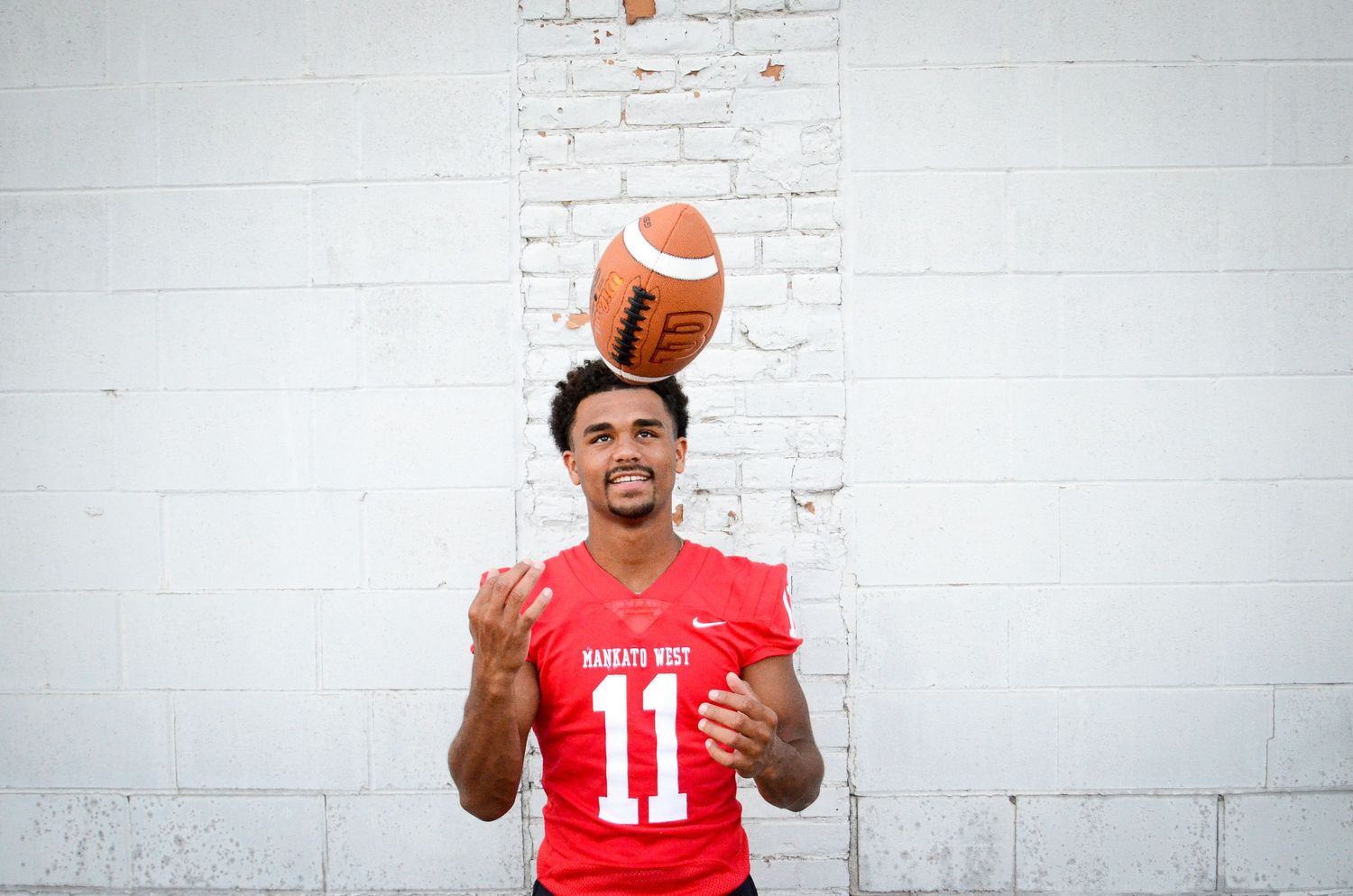 Male high school football player in red jersey tossing a football in the air against a white wall.