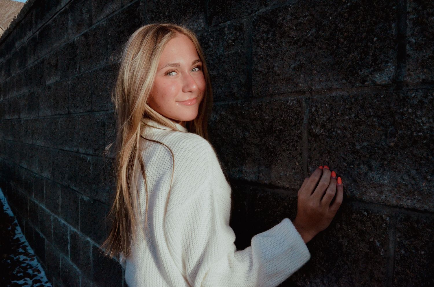 Young blonde woman in white coat smiling over shoulder against a stone wall outdoors.