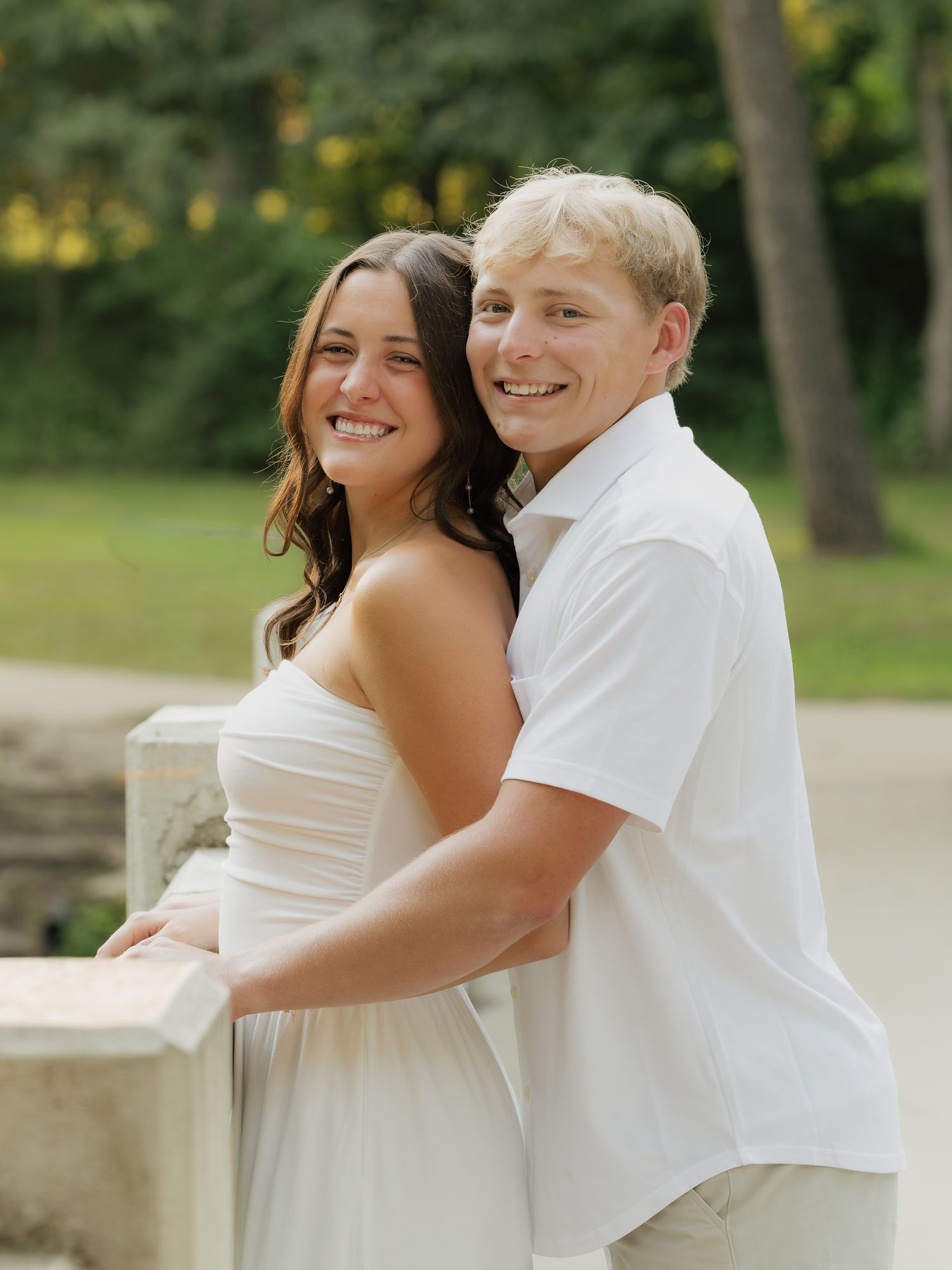 A smiling young couple in white outfits poses outdoors near a stone railing with lush green trees behind them.