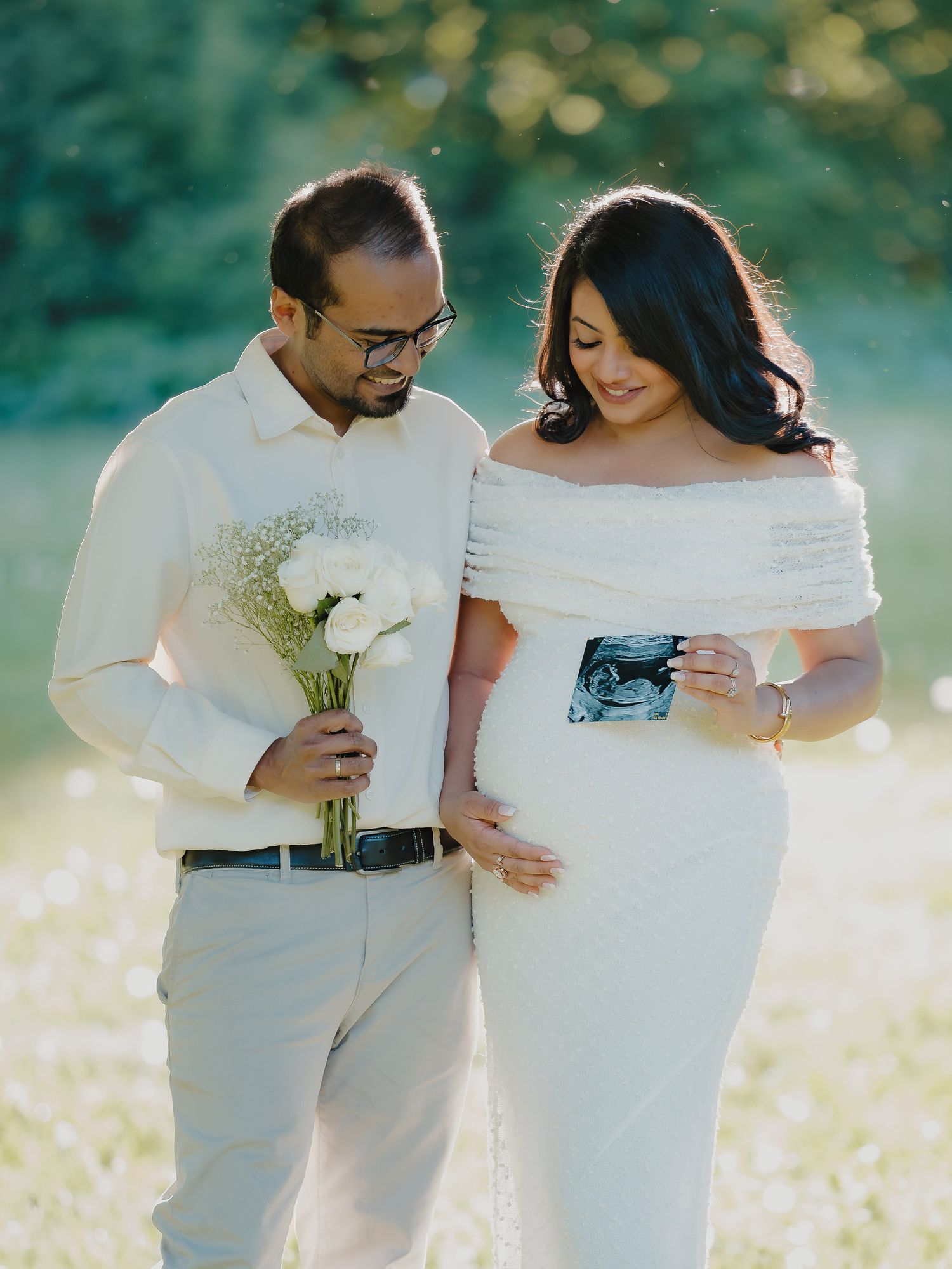 Expecting couple in white outfits holding ultrasound photo and flowers in sunny green meadow.
