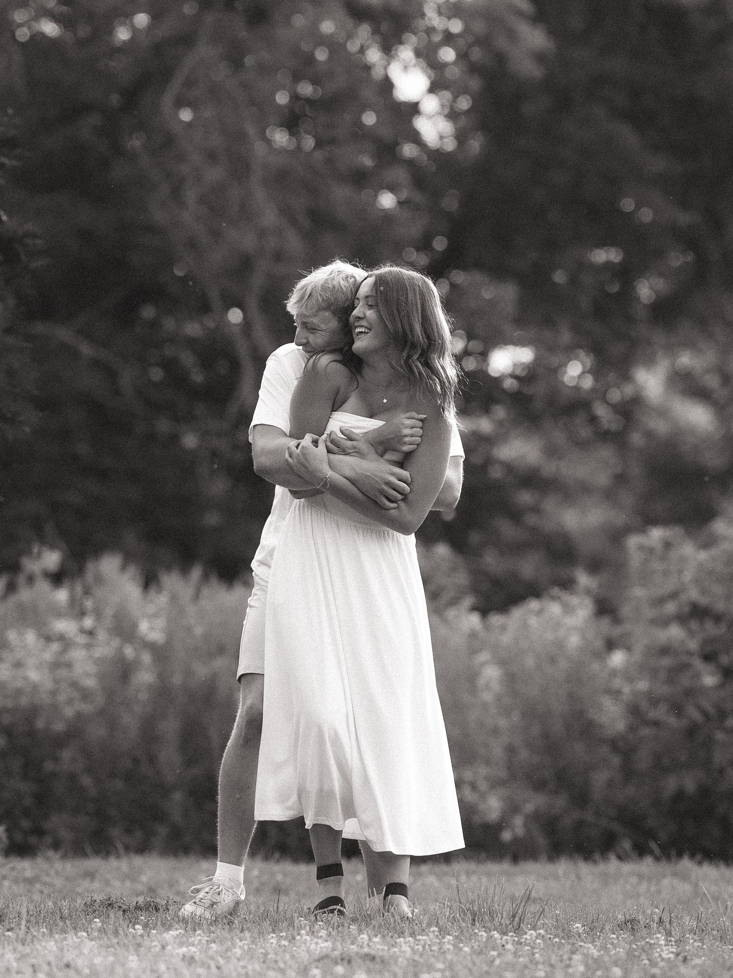 Black and white photo of a woman in a white dress embracing a child outdoors in a grassy field with trees.