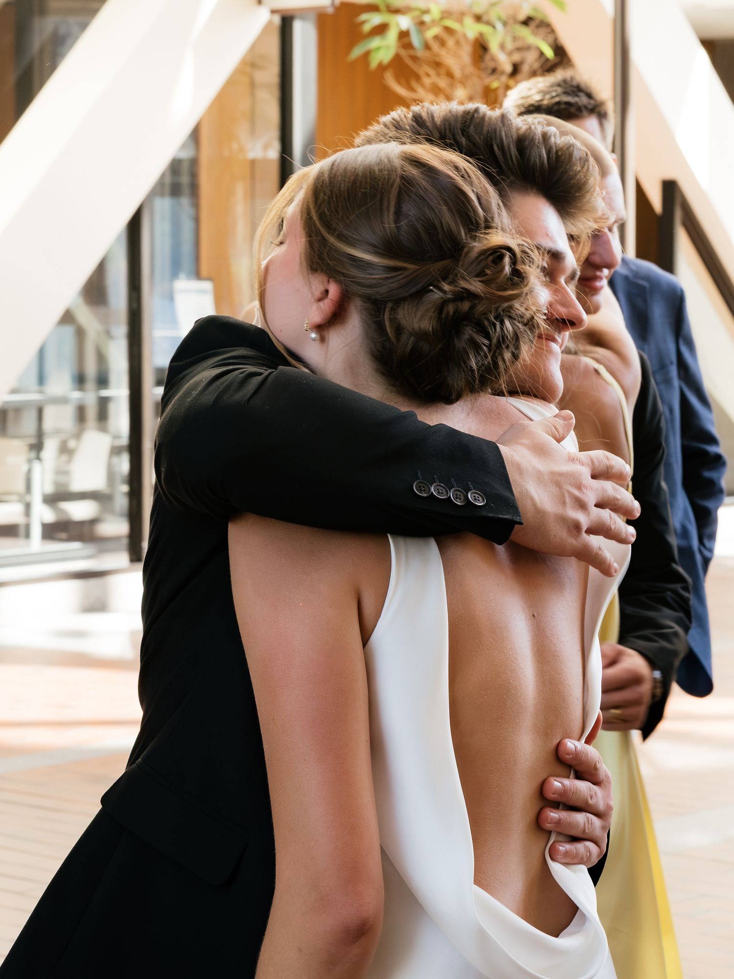 Two women share a warm embrace at a wedding, one in a white backless gown, surrounded by guests.