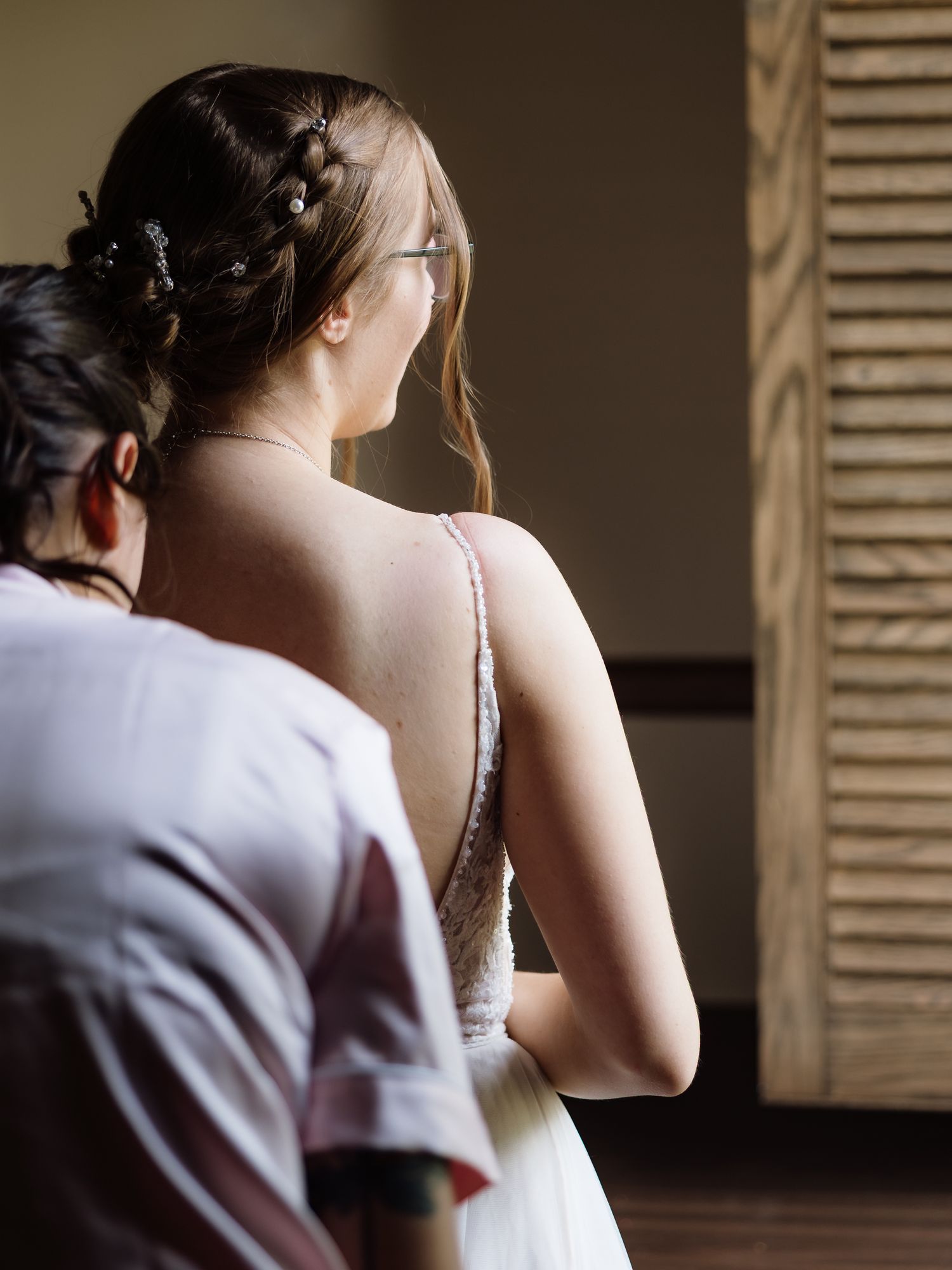 Bridesmaid helping bride with wedding gown in softly lit indoor setting.