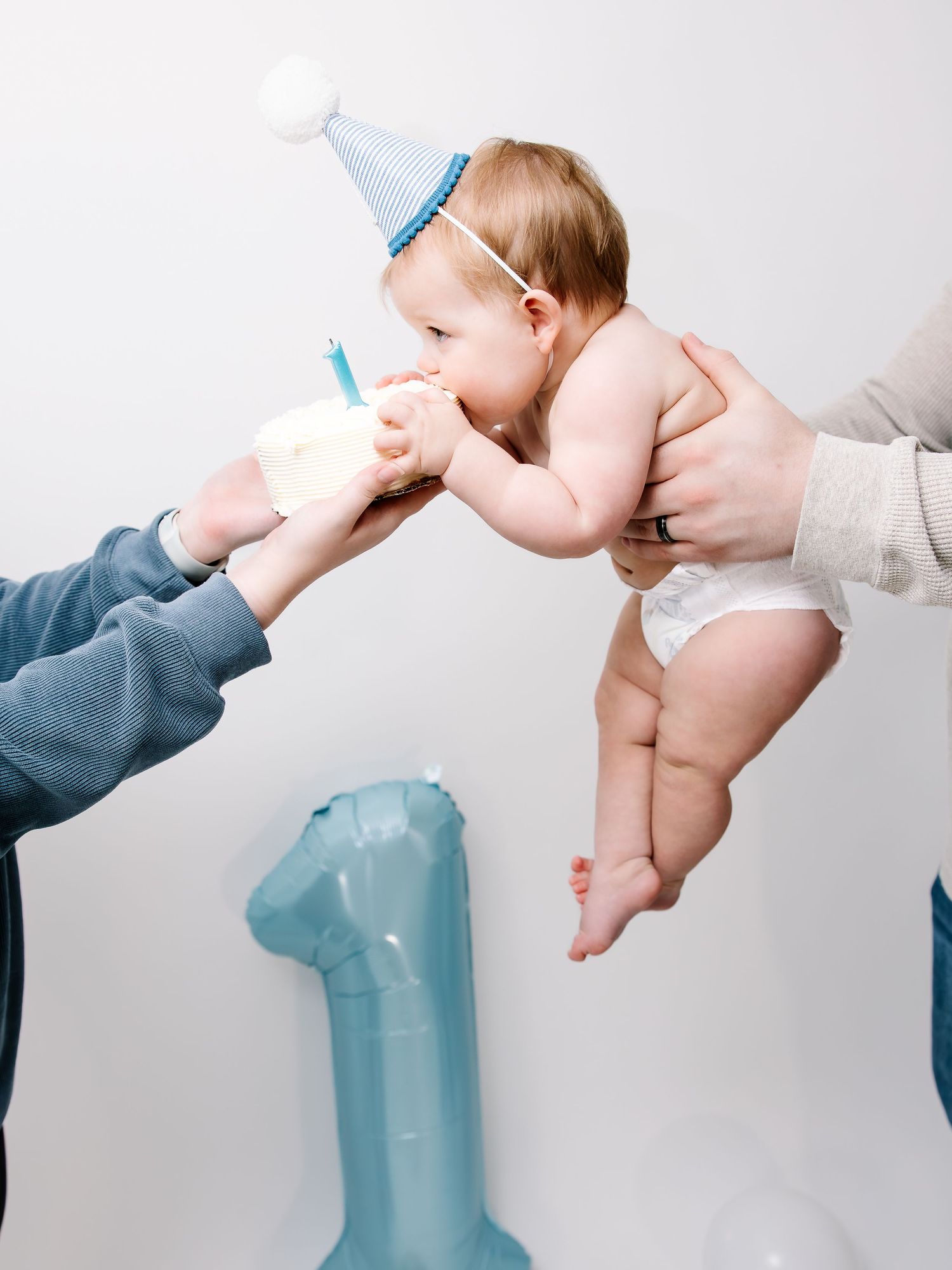 Baby in party hat eagerly eating first birthday cake while being held by parents.