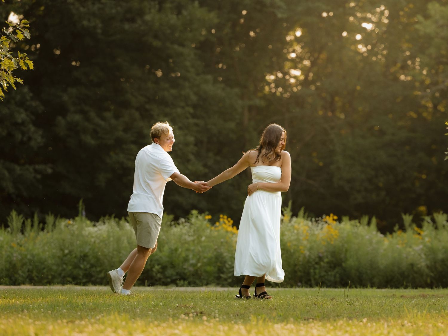 Couple holding hands and dancing playfully in a sunlit green park during golden hour.