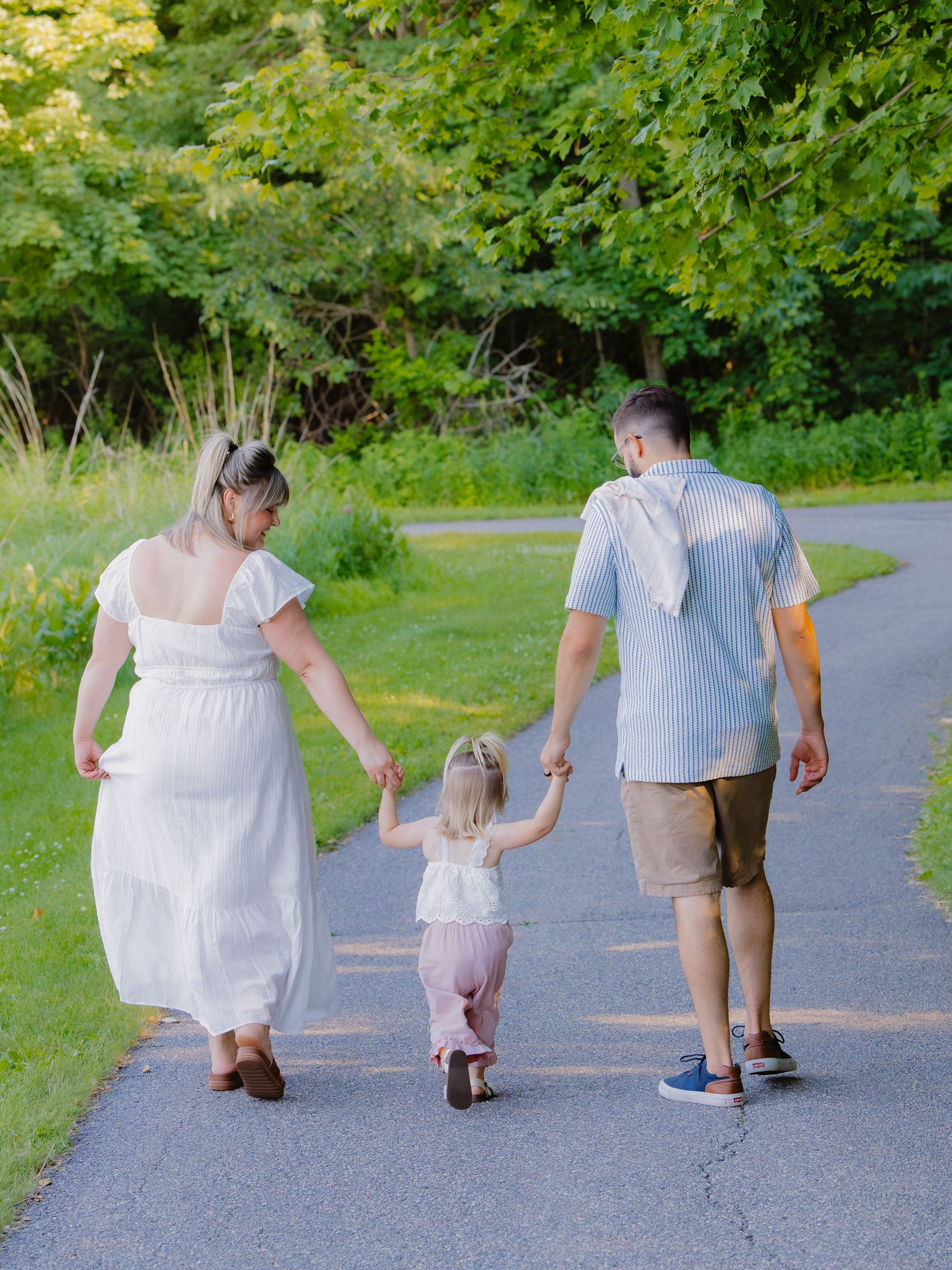 Family of three walking hand-in-hand down a tree-lined path in summer, viewed from behind.